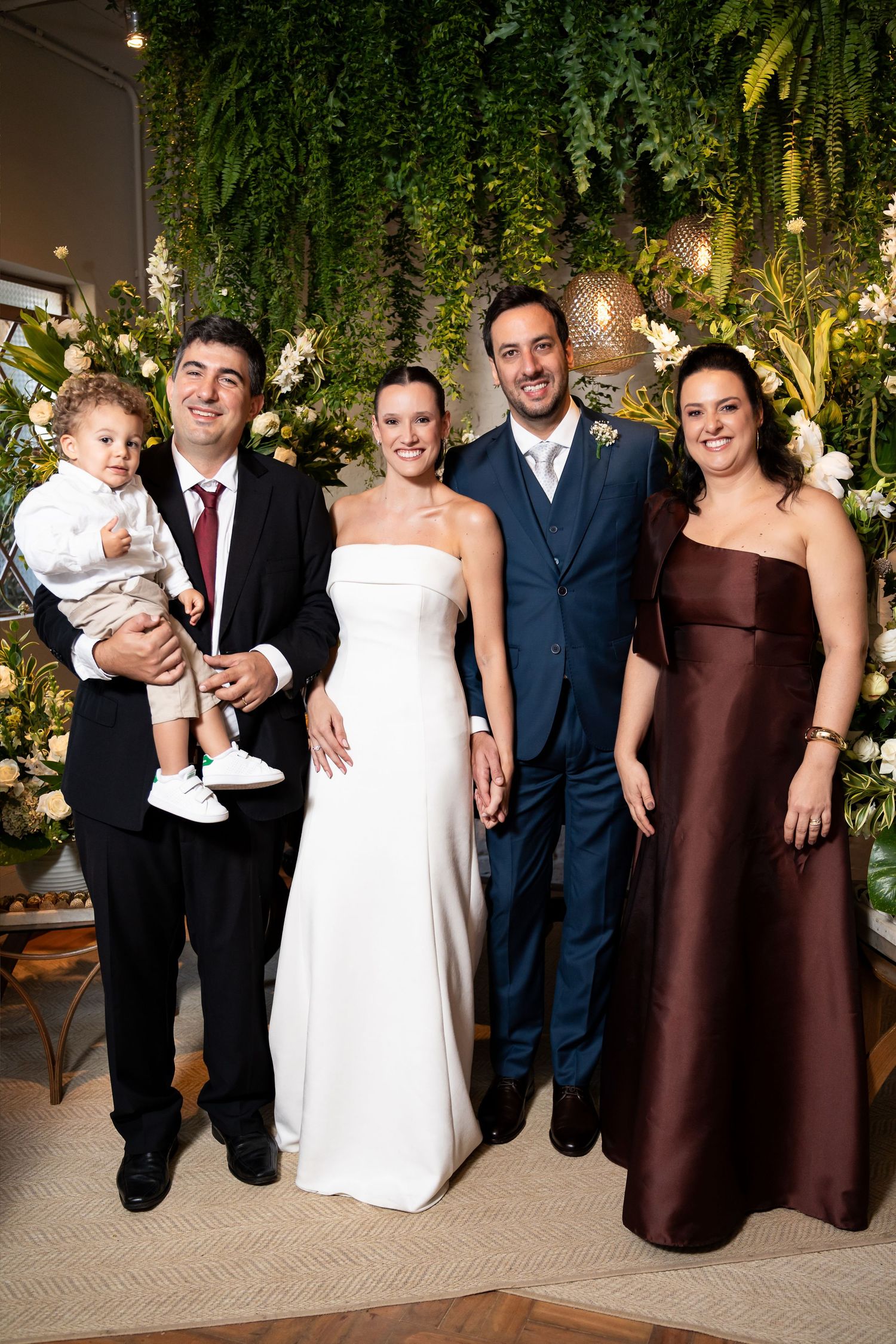 Wedding party posing with bride in white gown, groom in navy suit, family members in formal attire, lush greenery backdrop.
