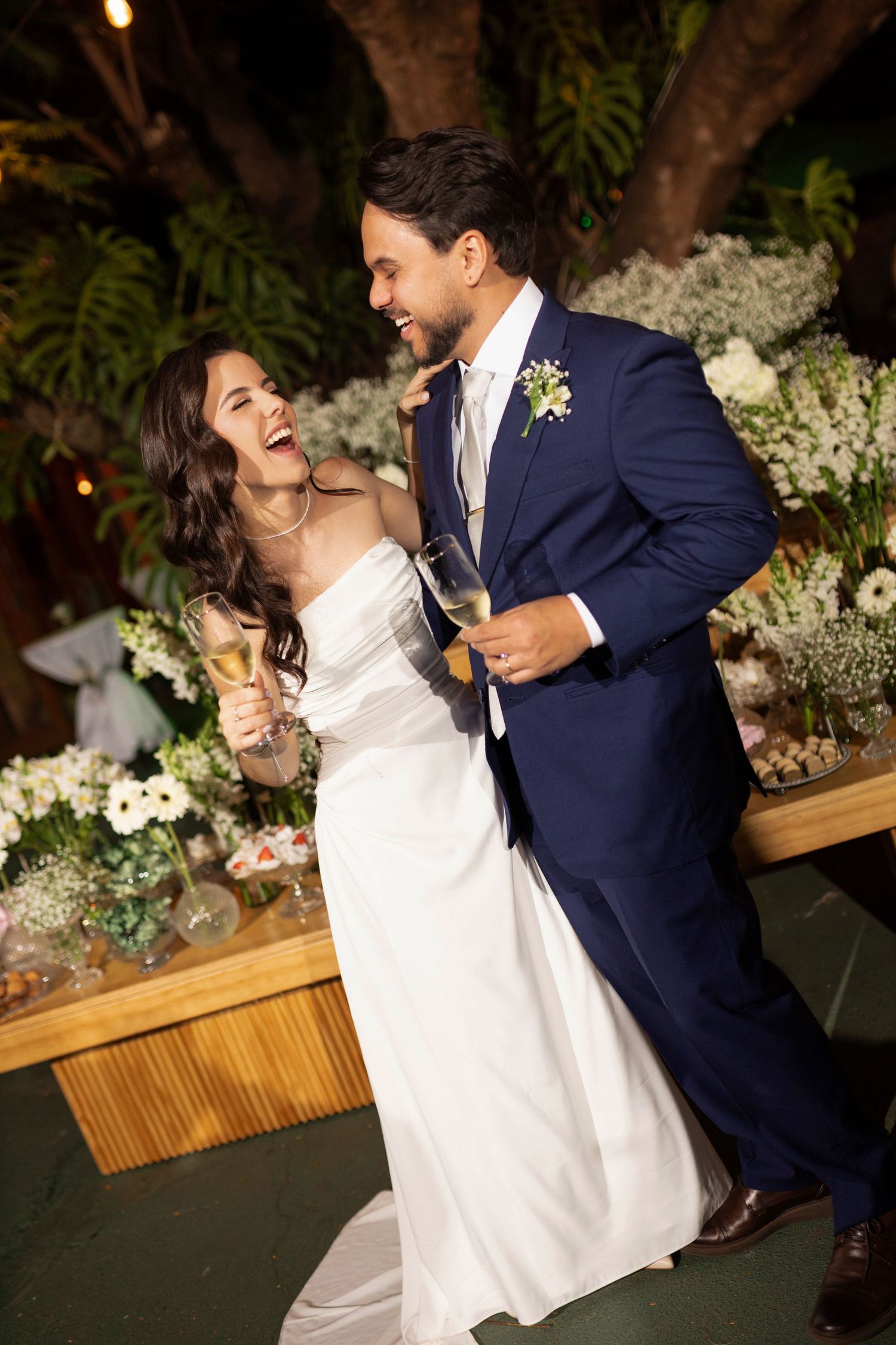 Joyful bride in white gown and groom in navy suit laughing and holding champagne glasses at their wedding reception.