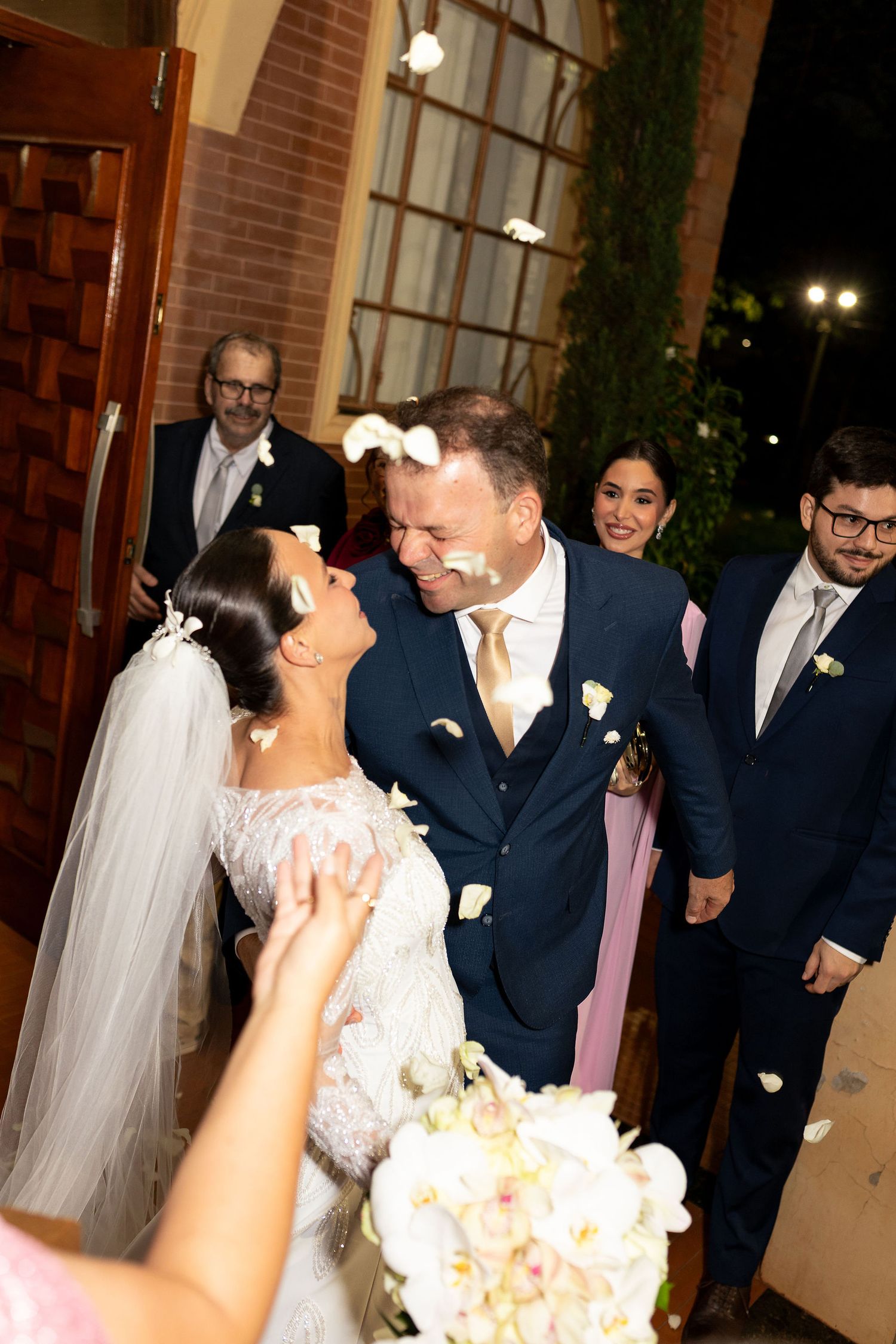 Newlyweds share a joyful laugh as guests toss white flower petals outside the wedding venue at night.