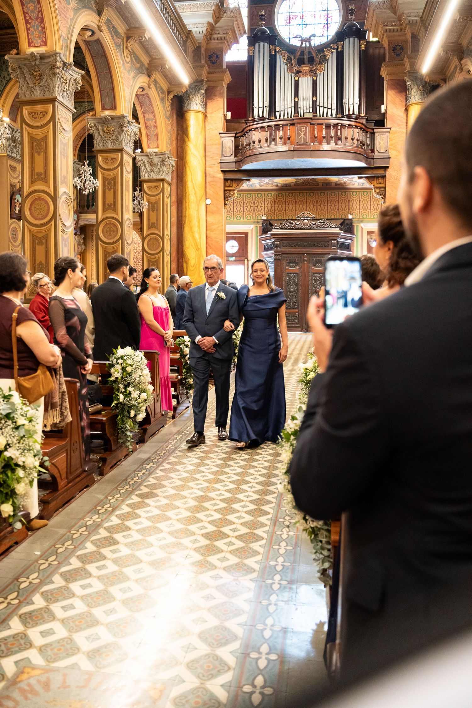 Elegant church wedding ceremony with couple walking down ornate aisle, pipe organ backdrop, guests seated in wooden pews.