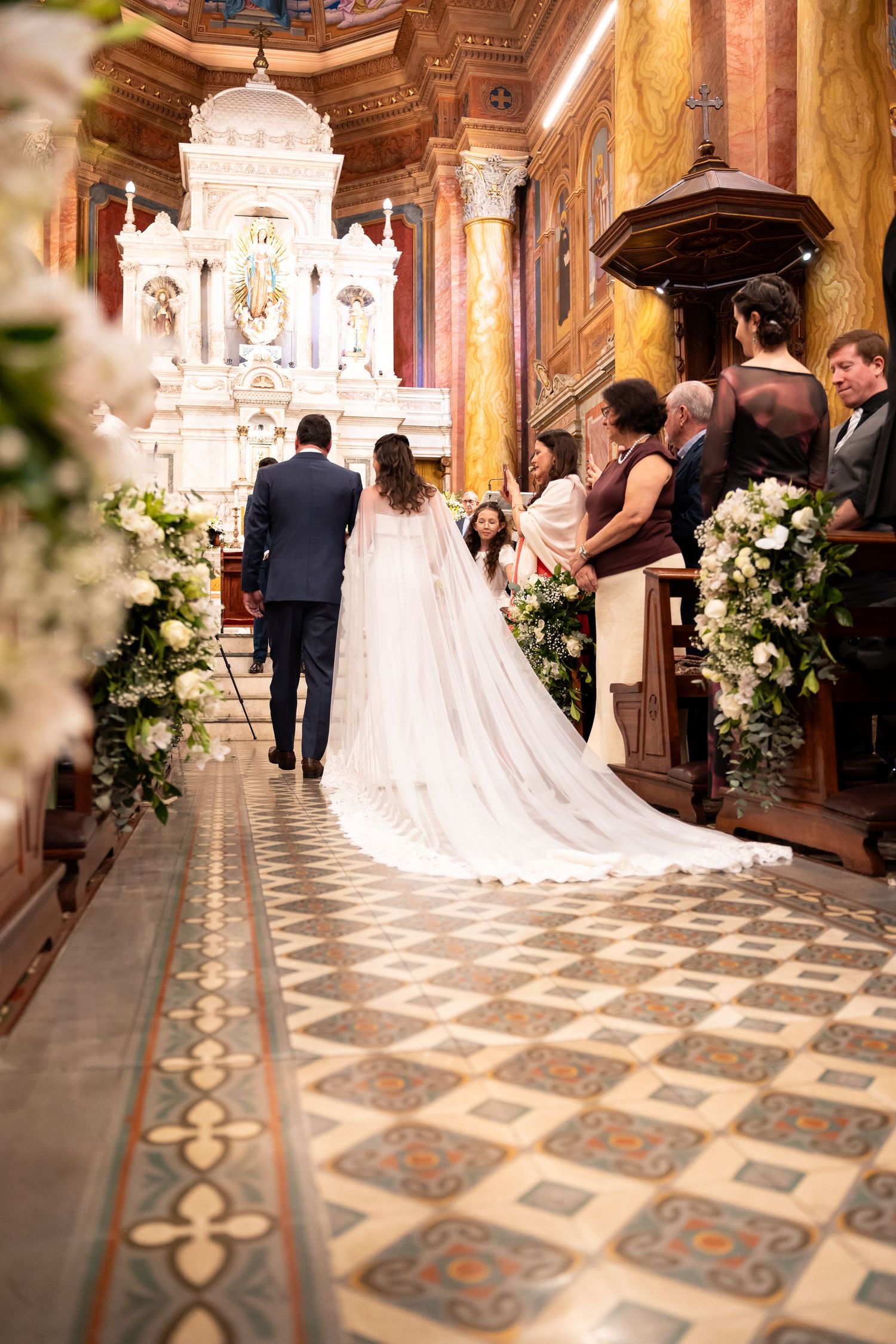 Bride and groom walking down ornate church aisle with white floral arrangements and decorative tile floor.