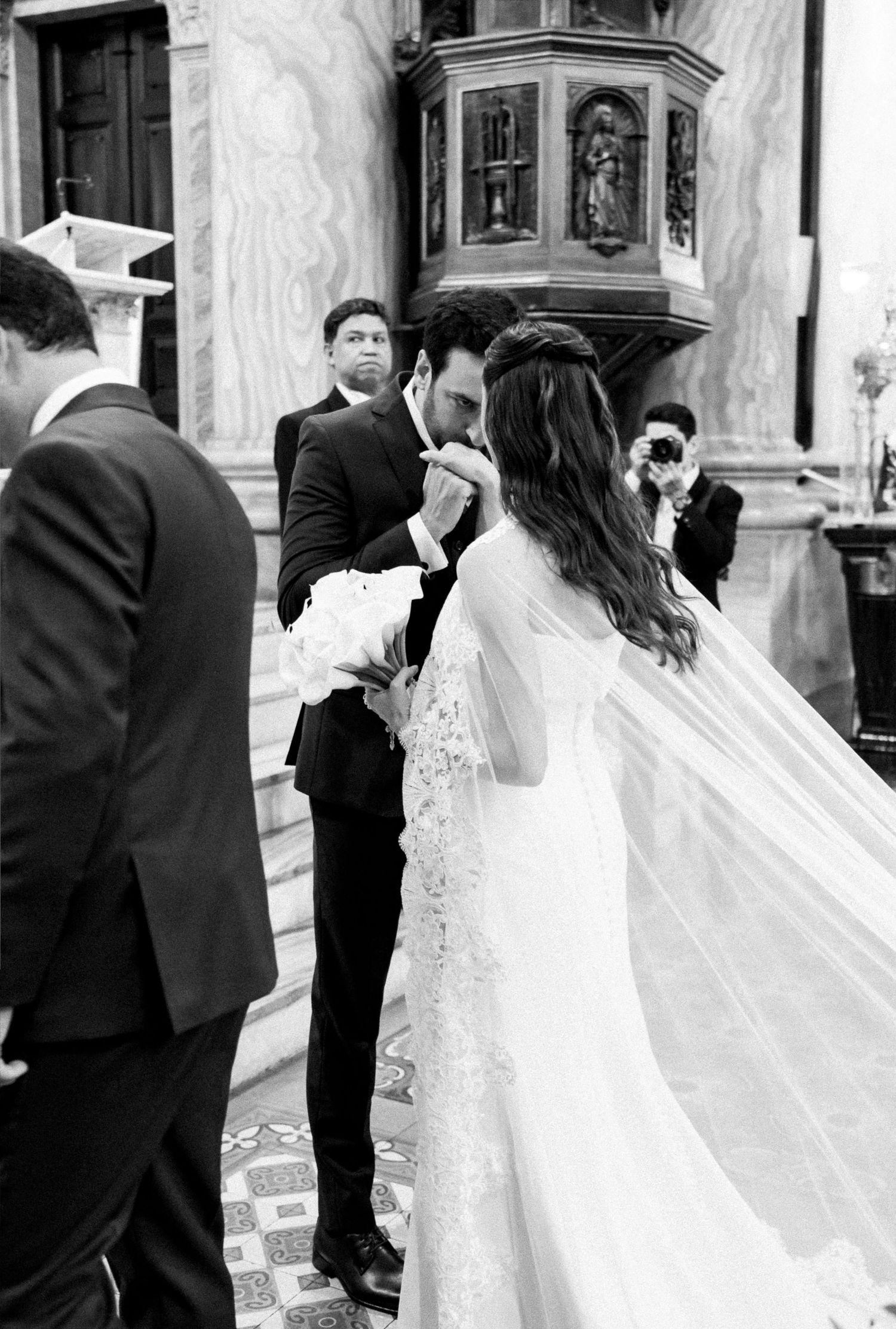 Groom kissing bride's hand at church altar in black and white wedding photo with veil flowing.