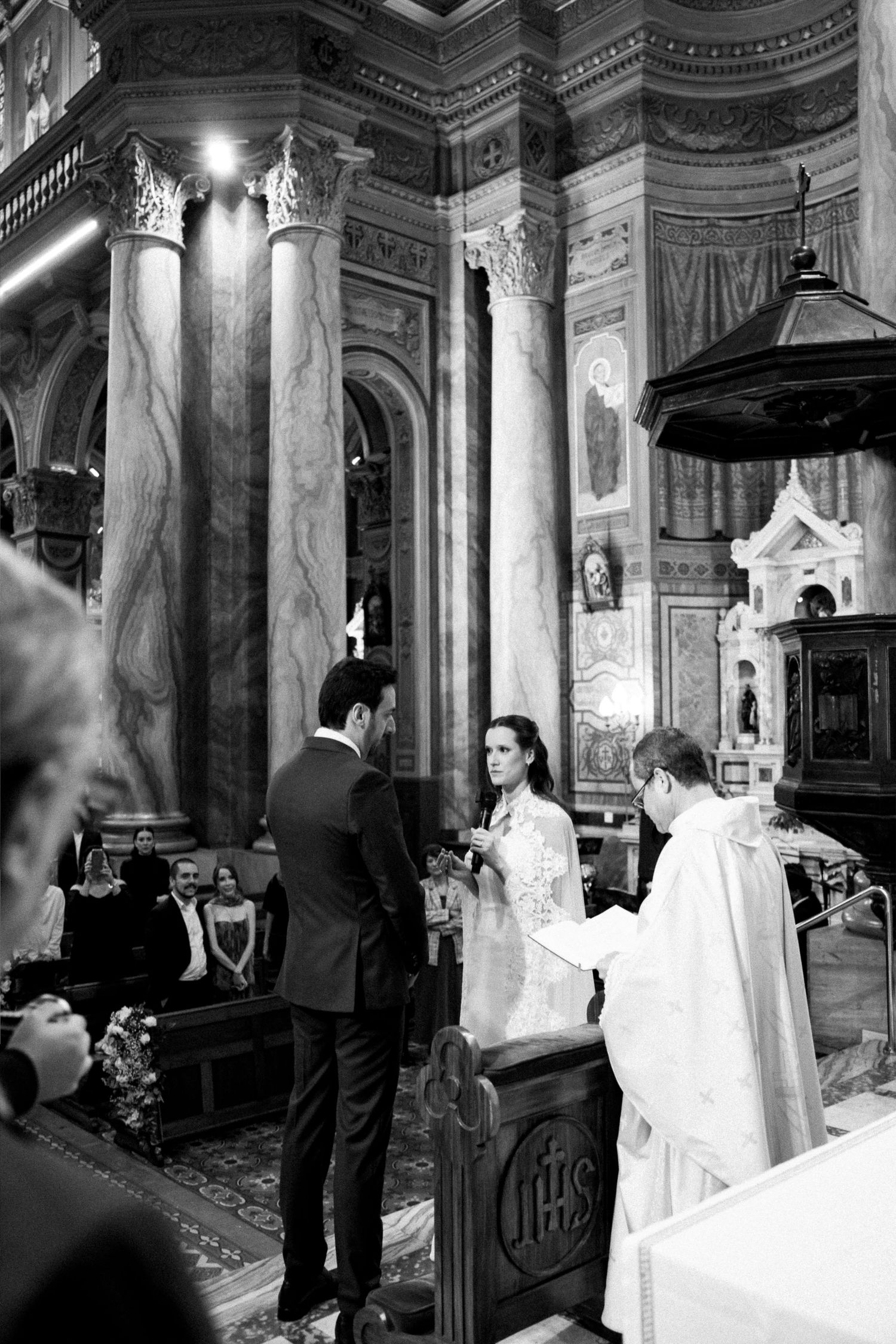 Black and white photo of a couple exchanging vows before a priest in an ornate church during a wedding ceremony.