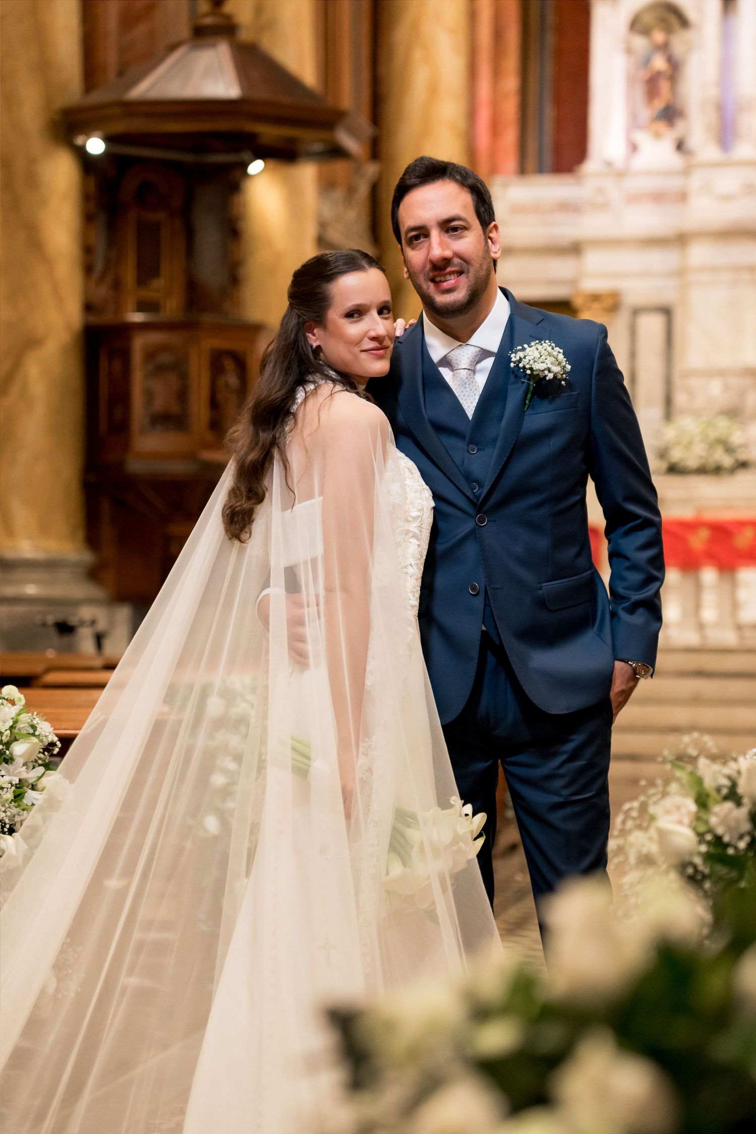 Bride in white gown with veil and groom in blue suit smiling together inside a church on their wedding day.
