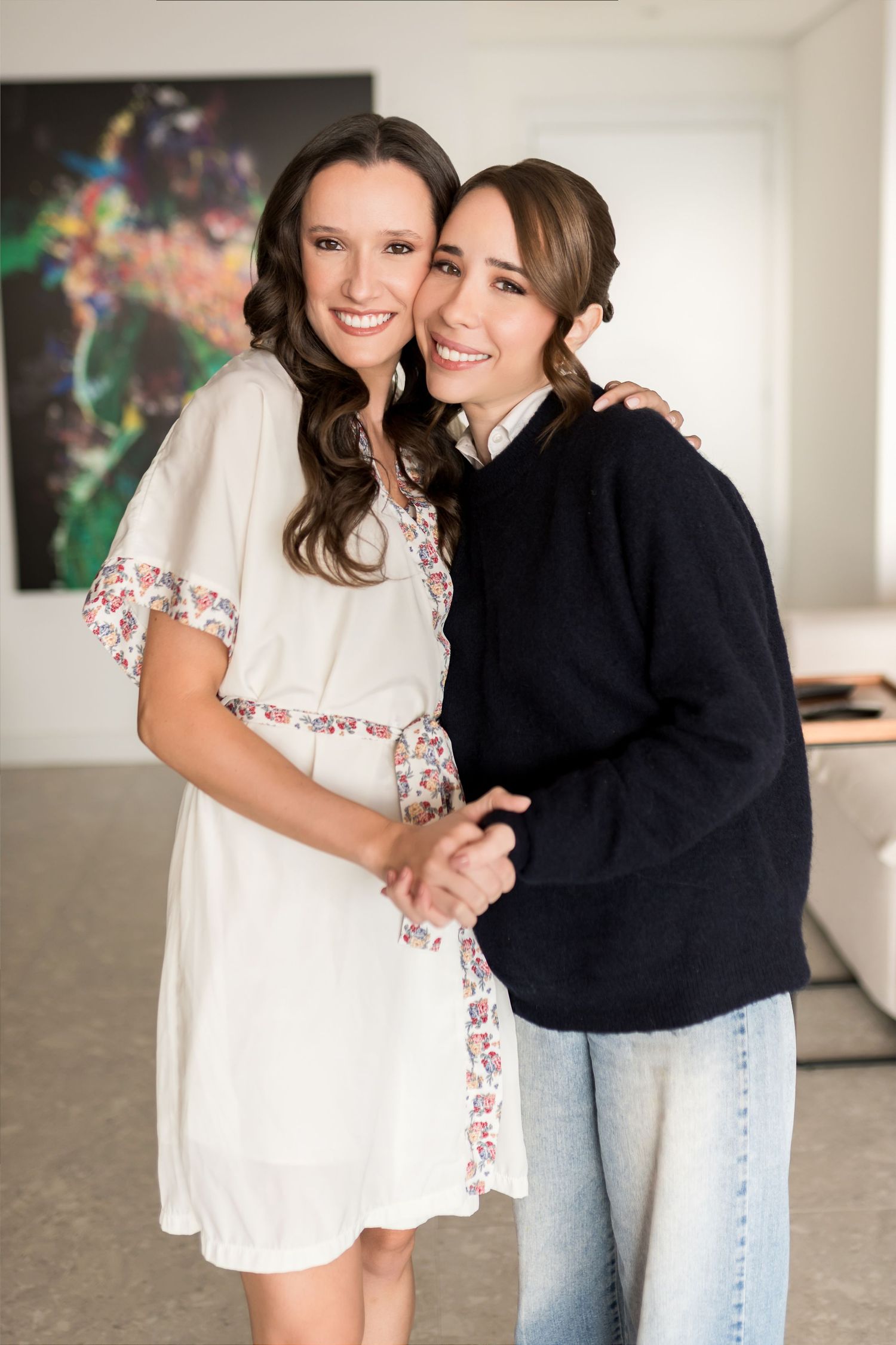 Two smiling women embrace indoors, one in a white floral dress and one in a navy sweater with jeans.