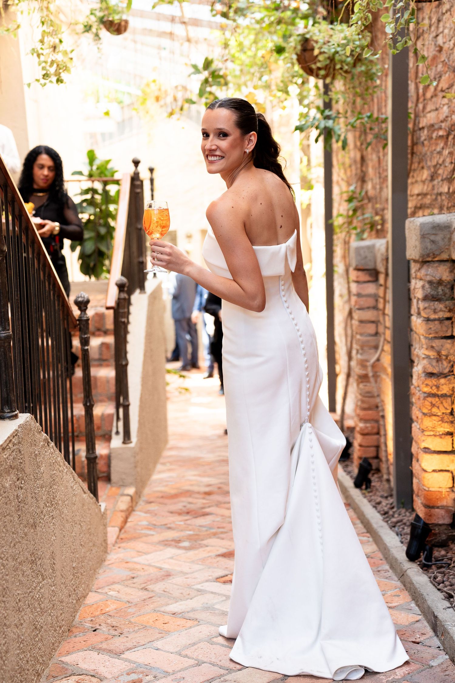 Bride in elegant white backless gown holding champagne flute, smiling over shoulder in outdoor garden setting.