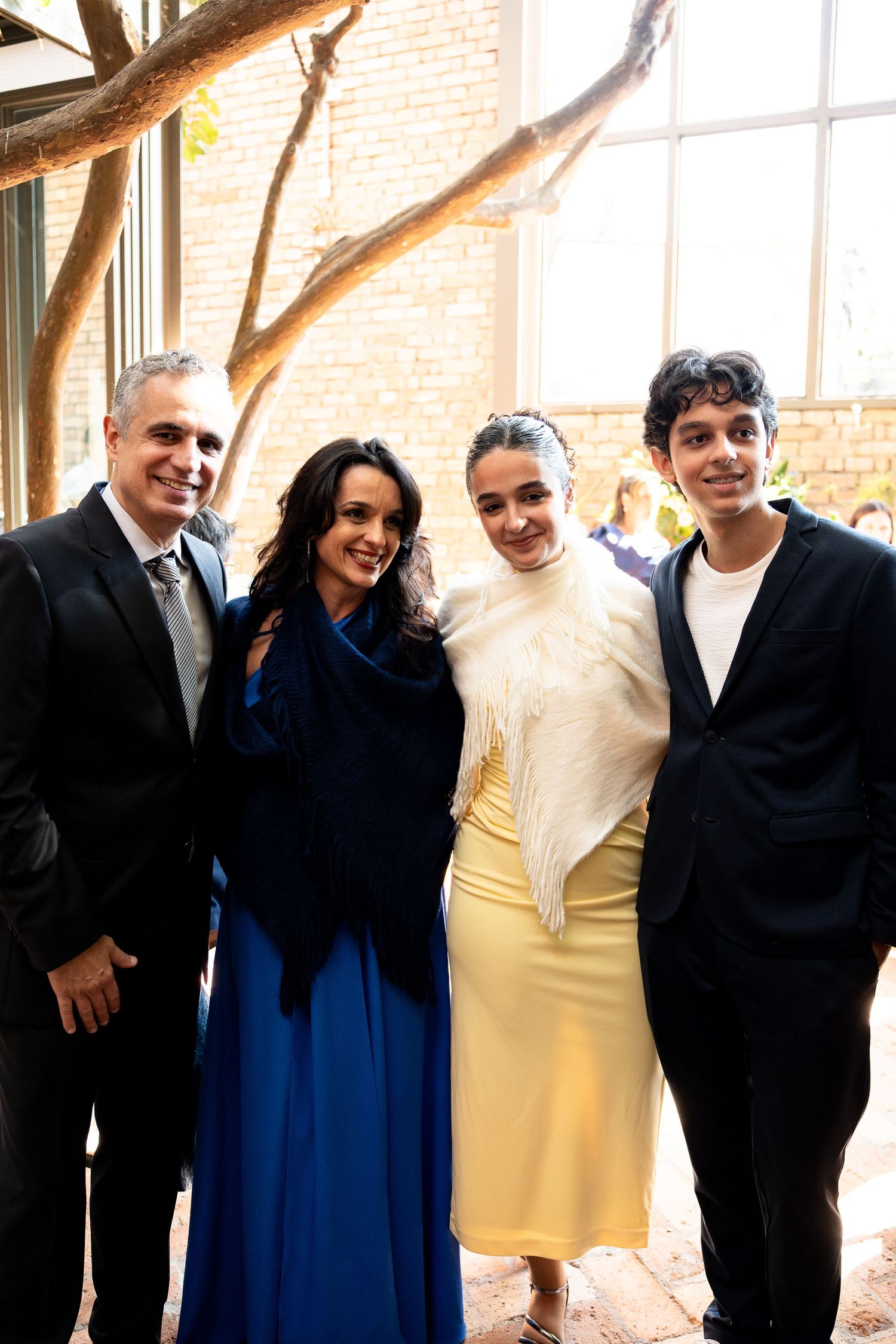 Four elegantly dressed people posing together indoors at a formal event near a brick wall and tree branches.
