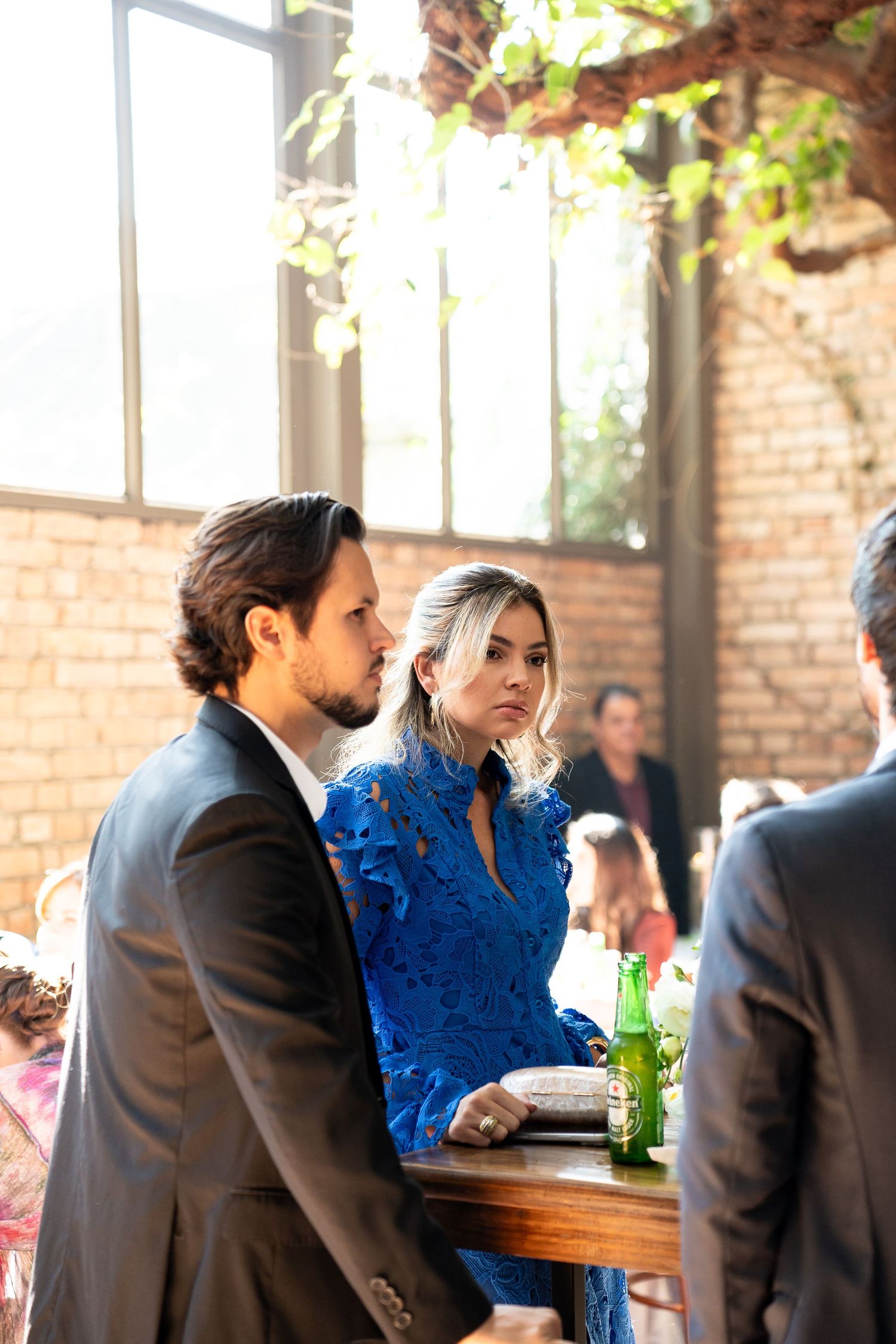 Couple in formal attire at an indoor event, woman in blue dress with green beer bottle on wooden table.