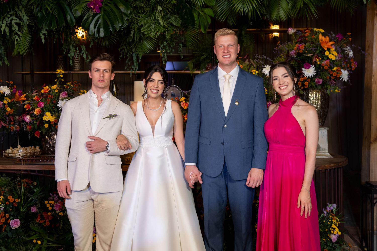 Wedding party of four posing indoors with tropical floral decor, featuring bride in white gown and guests in formal attire.