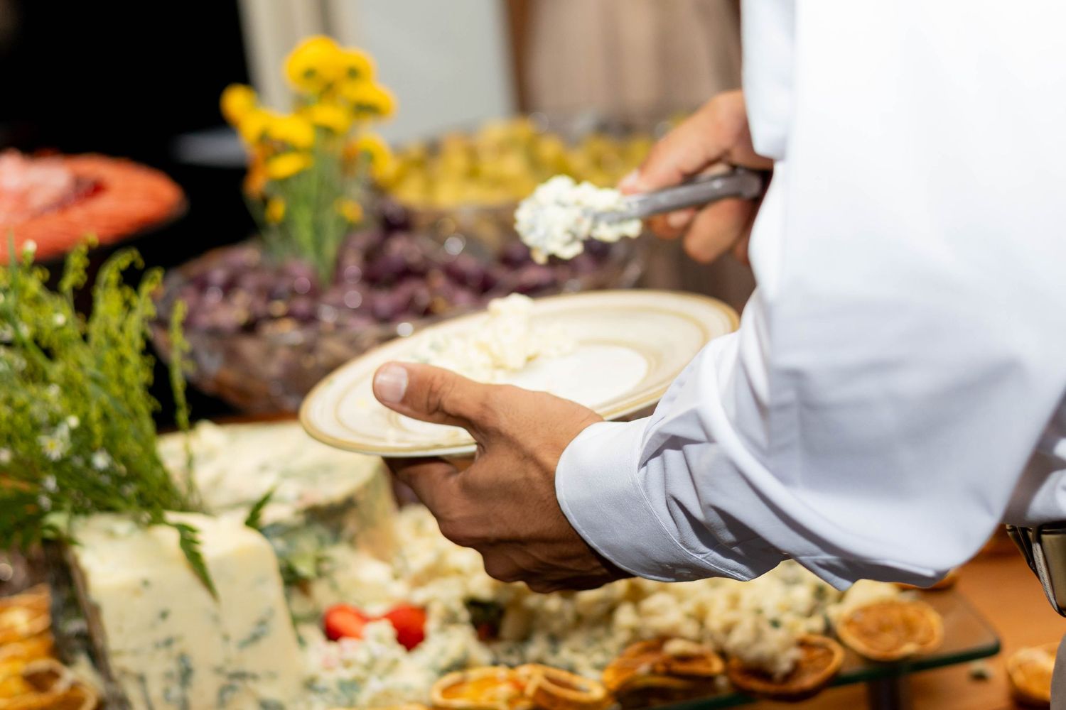 Catering chef serving creamy cheese spread at an elegant buffet table with fresh flowers and appetizers.
