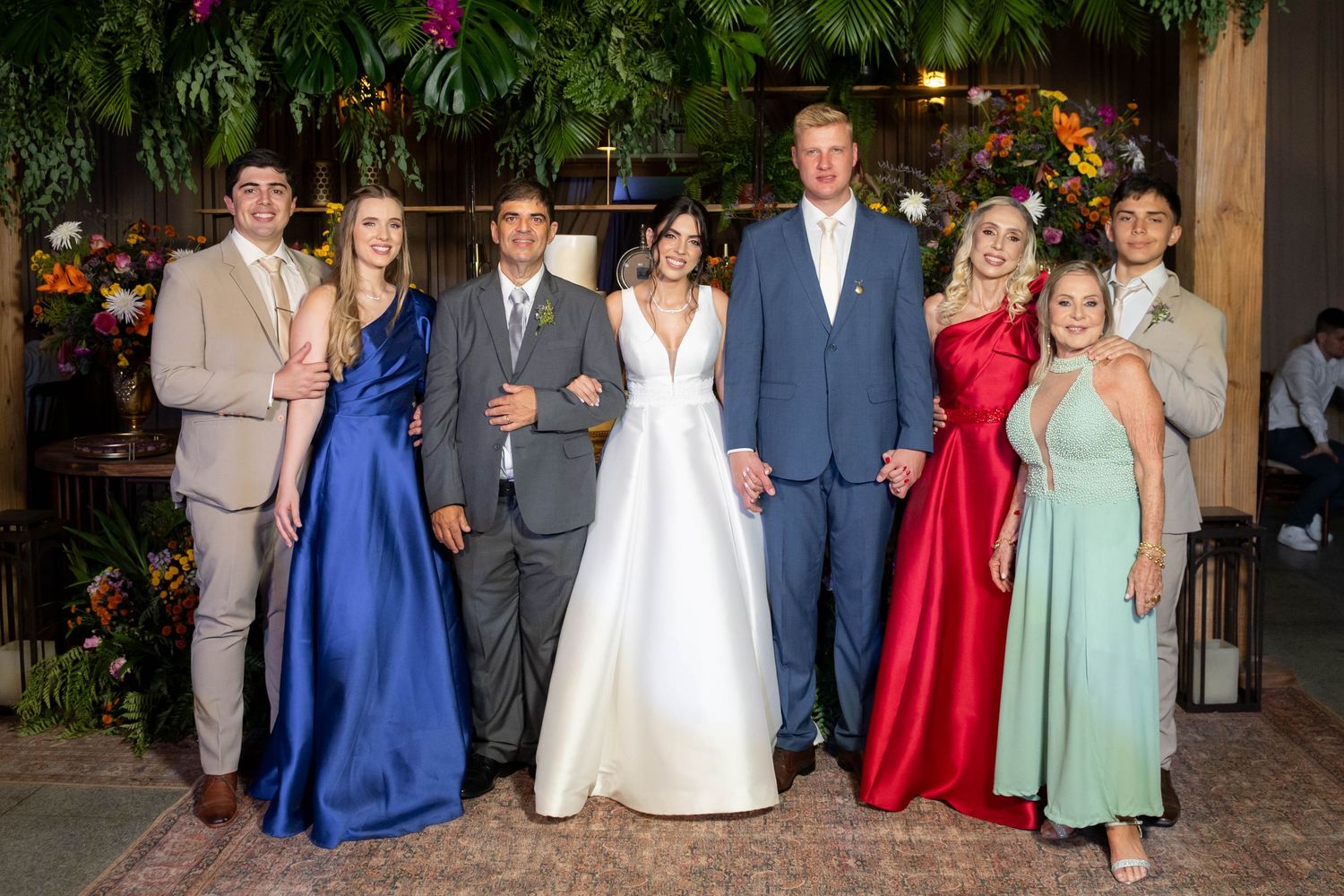 Wedding party of seven posing together, bride in white gown flanked by family in formal attire amid lush floral decor.