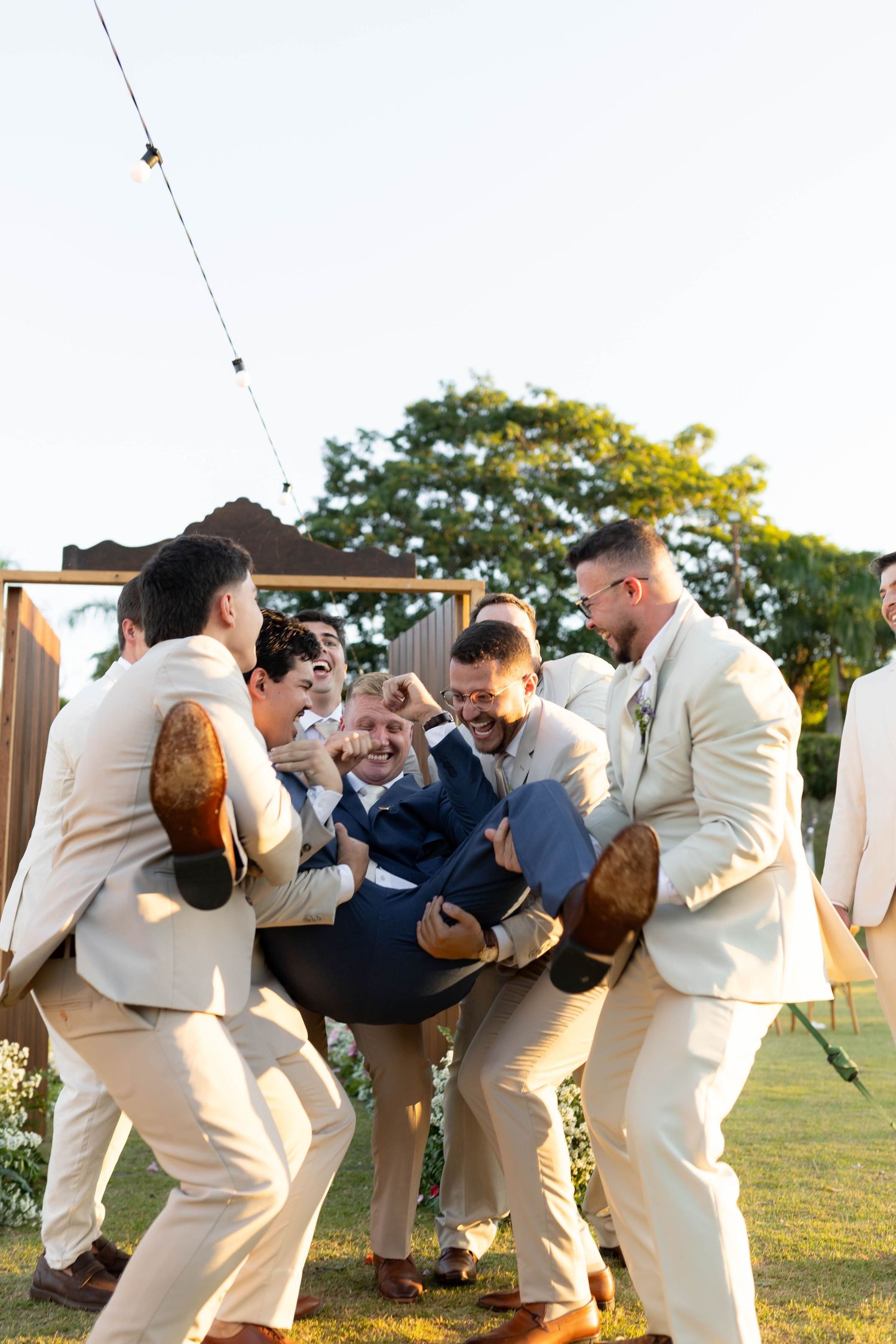 Groomsmen in tan suits joyfully lift the groom in a navy suit during an outdoor wedding celebration.