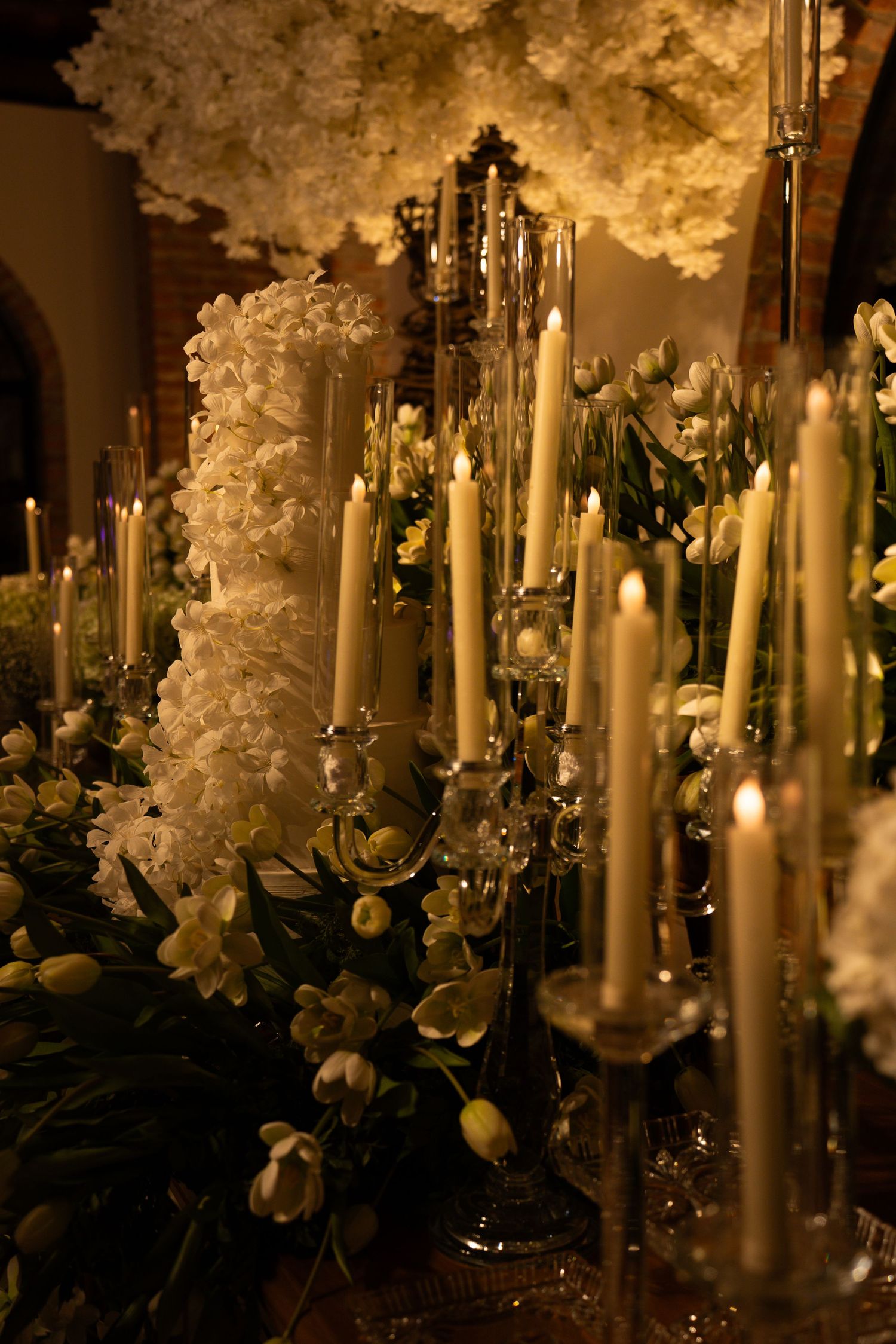 Elegant wedding decor with tall white candles, crystal candelabras, and white floral arrangements in warm golden light.