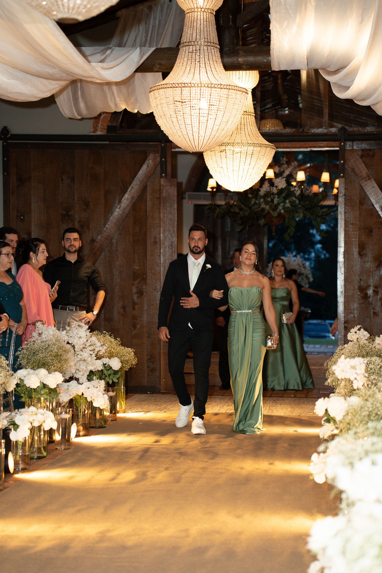 A couple walks down a floral-lined wedding aisle in a rustic barn venue adorned with elegant chandeliers.