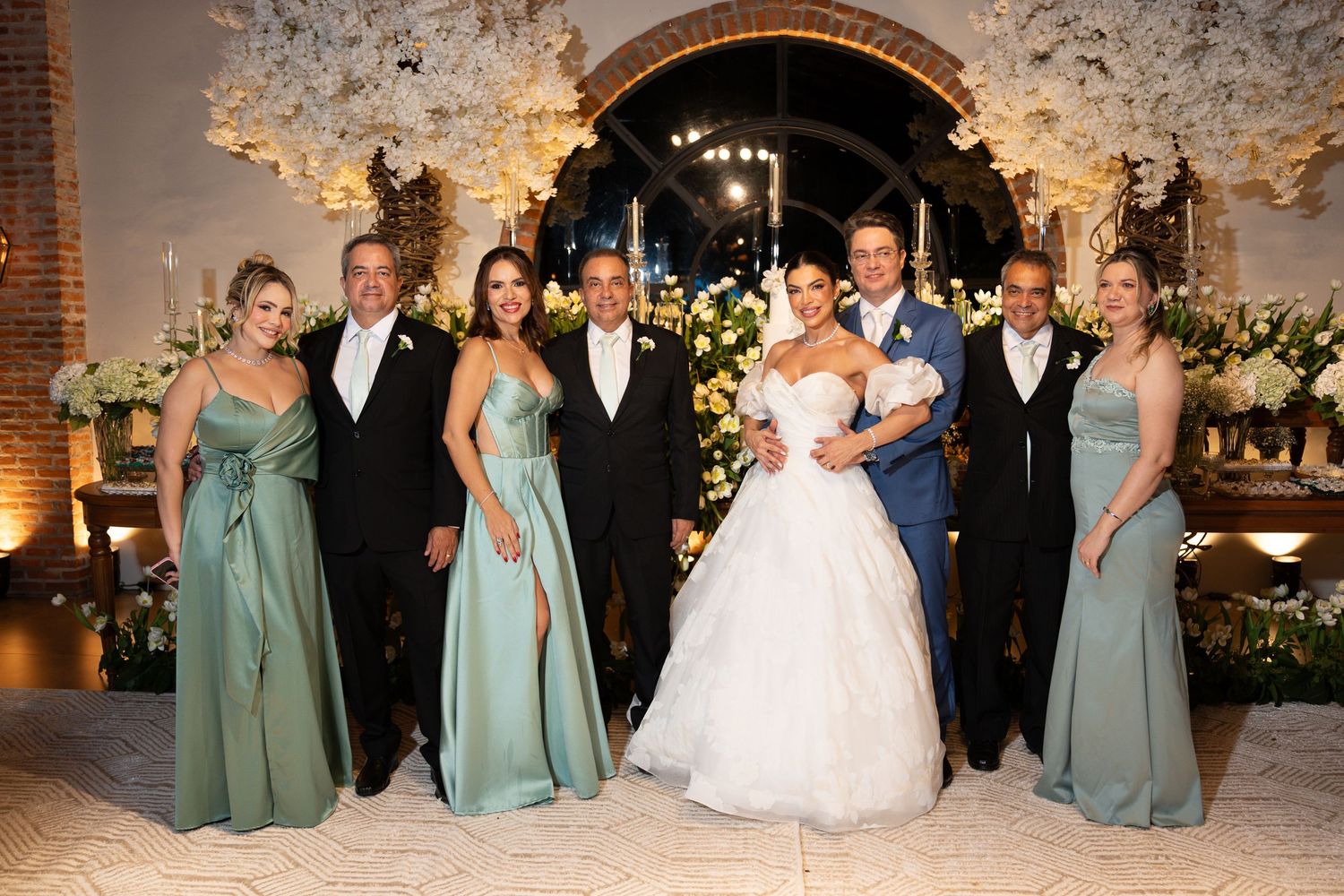 Wedding party poses together in elegant venue with floral arch, bride in white gown, bridesmaids in sage green dresses.