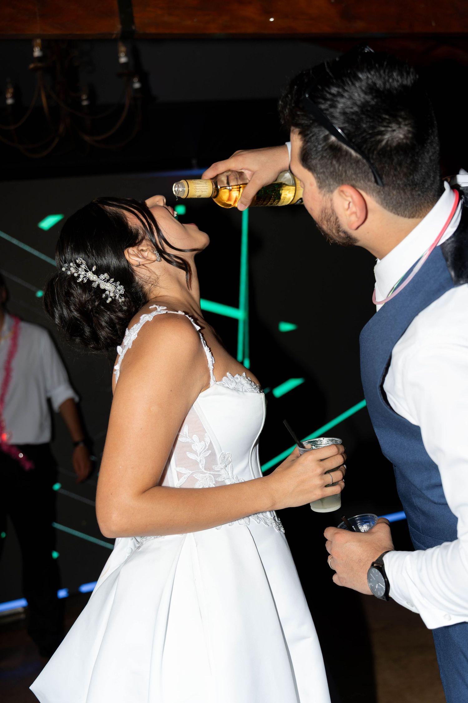 Bride and groom celebrating at wedding reception, groom pouring champagne bottle while bride holds a drink on the dance floor.