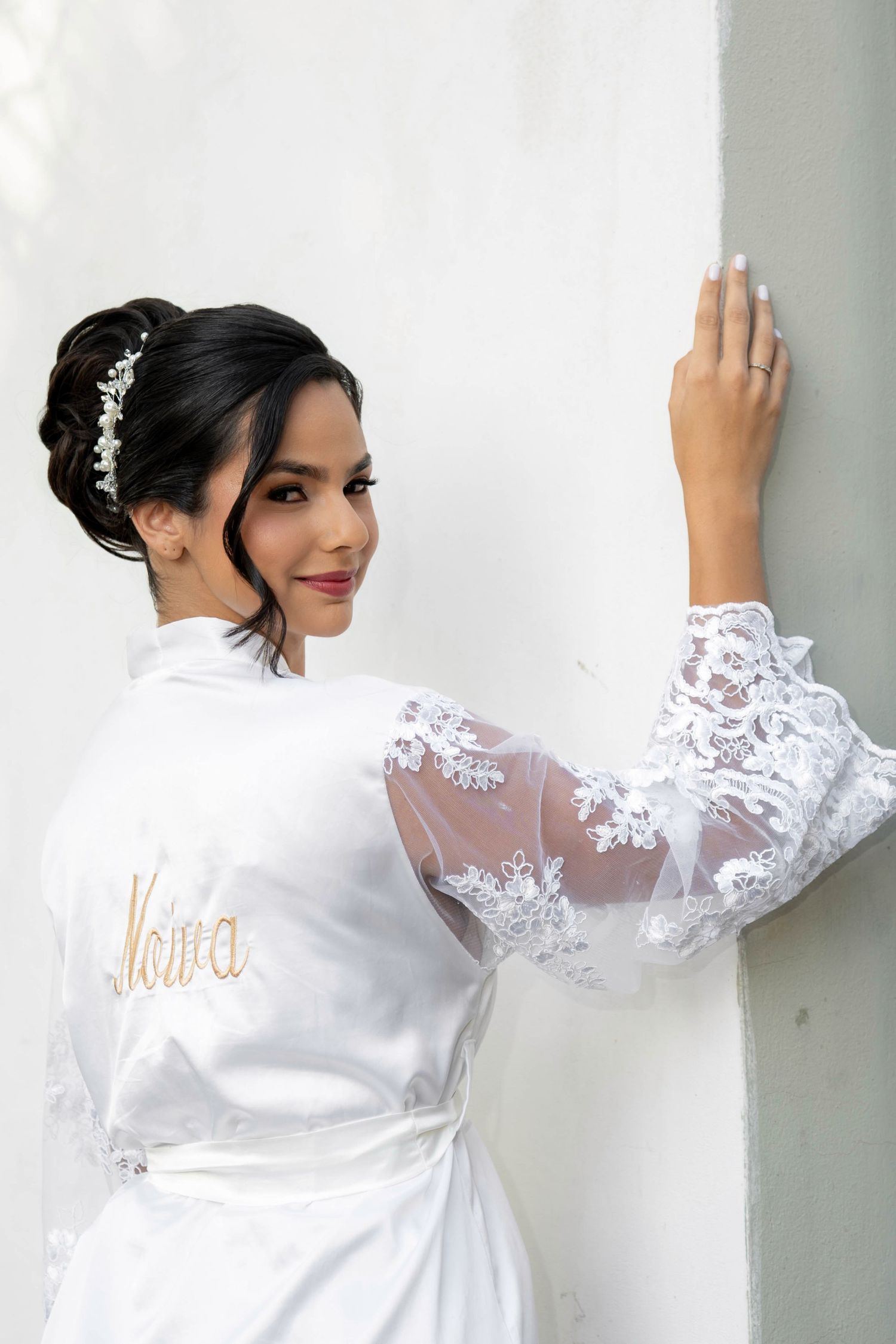 Bride in white lace robe with gold embroidered name, elegant updo with hair accessory, posing against white wall.