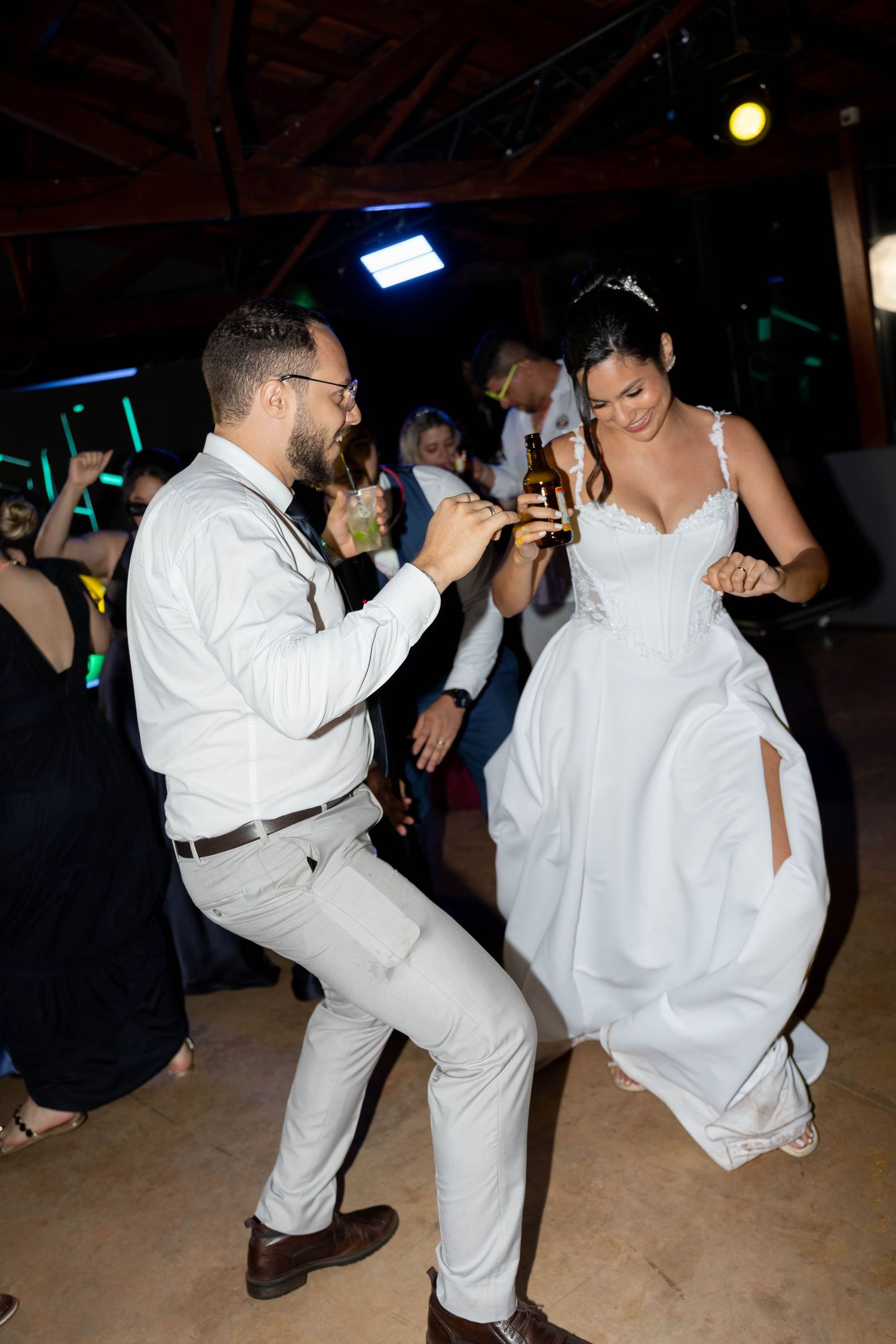 Bride and groom dancing joyfully at wedding reception, holding drinks, surrounded by guests in dimly lit venue.