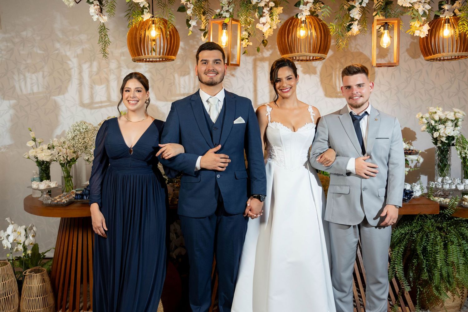 Wedding party of four posing elegantly in a decorated venue with floral arrangements and warm lighting.