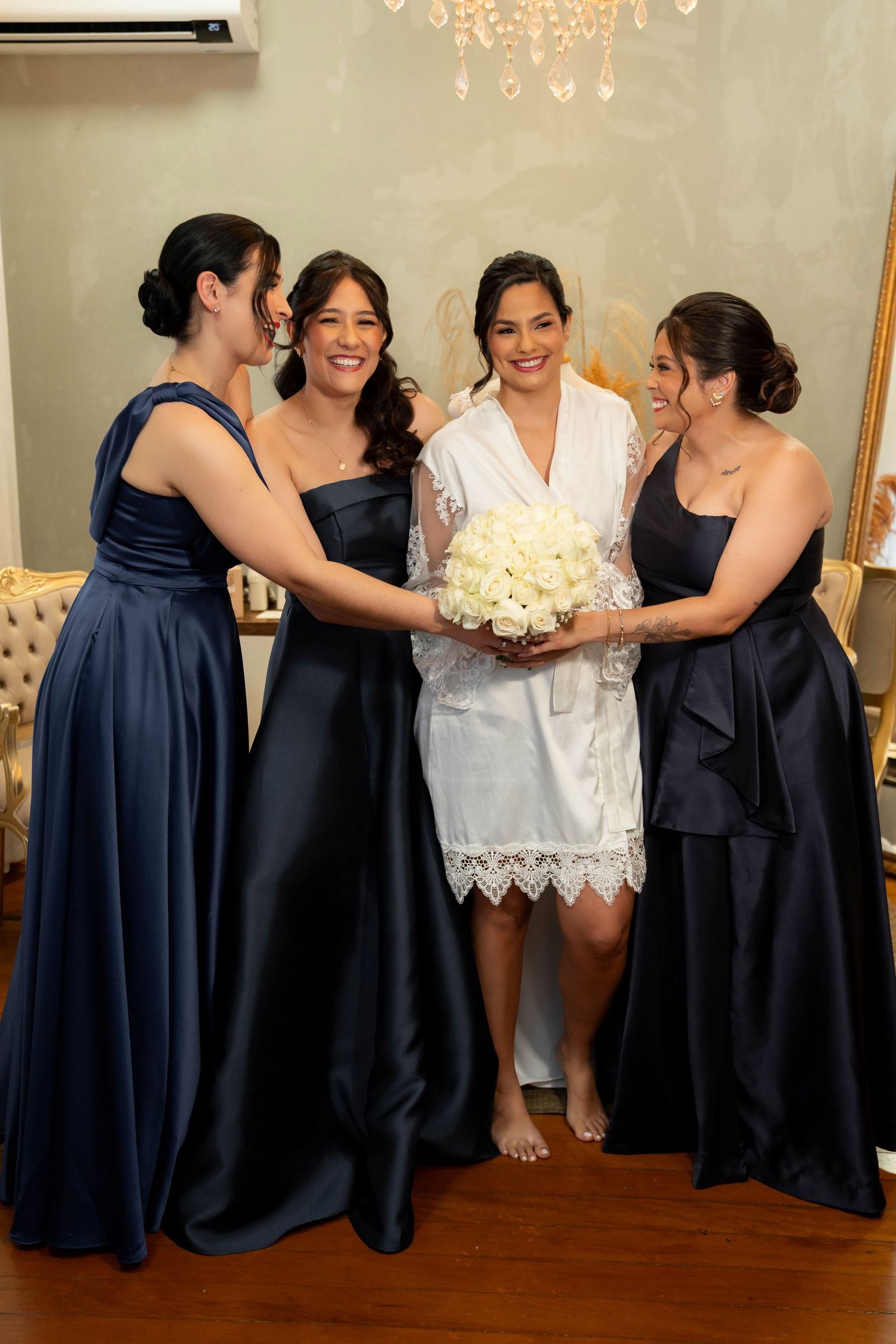 Bride in white lace robe holds white bouquet surrounded by three bridesmaids in navy blue gowns in elegant room.