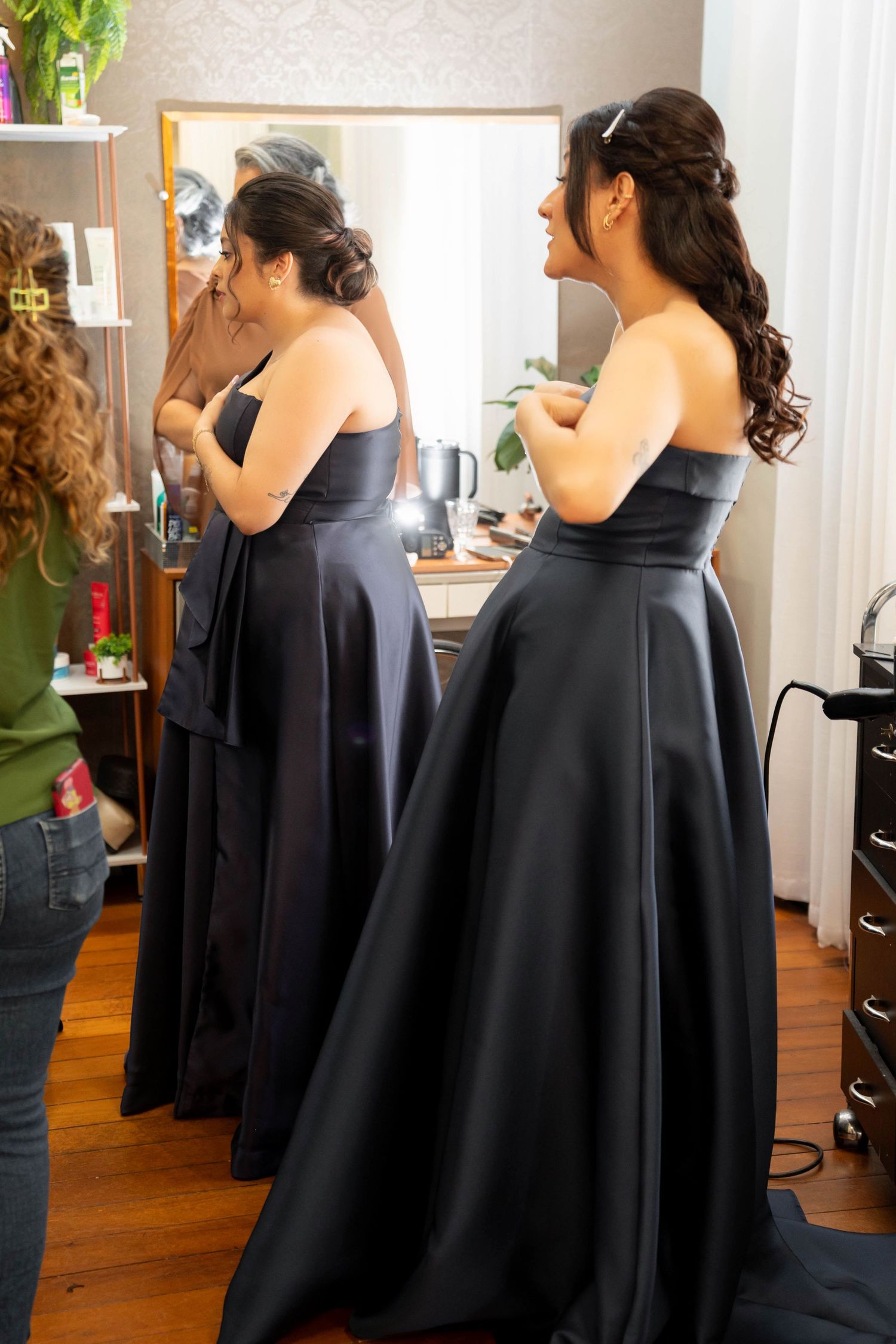 Two women in elegant black strapless gowns getting ready in a dressing room with mirror lights.