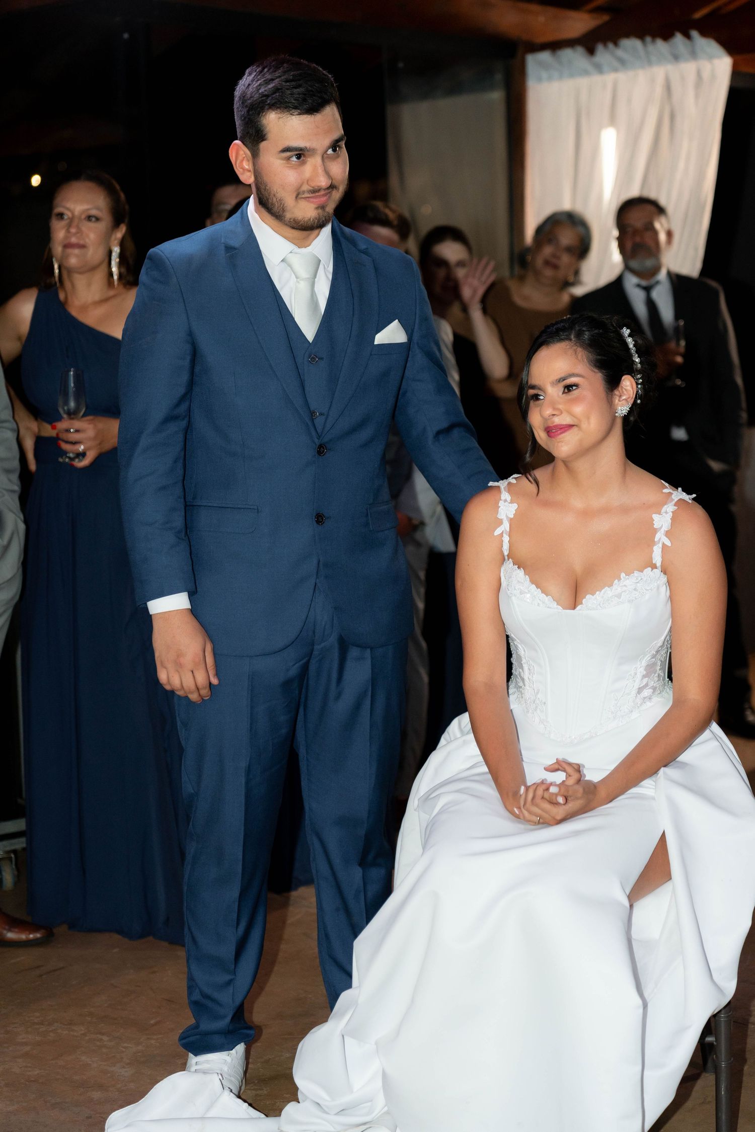 Bride in white lace gown sits beside groom in blue suit at wedding reception with guests in background.