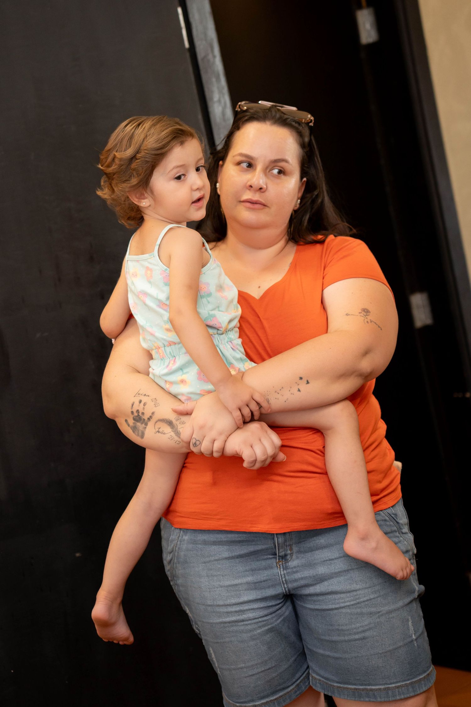 Woman in orange shirt holding a young toddler girl, both posing indoors, woman has tattoos on her arms.