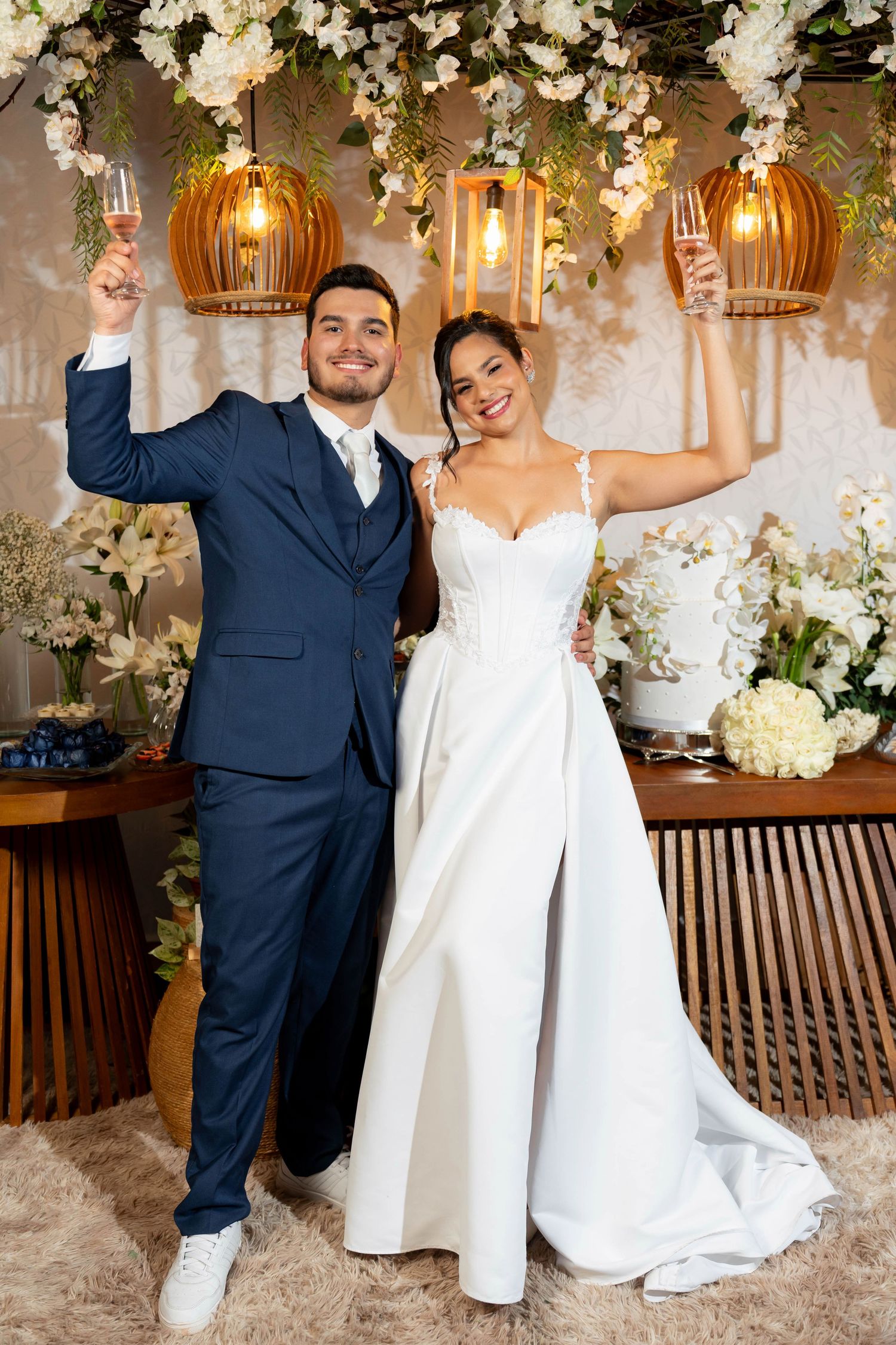 Bride and groom toast champagne in elegant wedding attire, surrounded by floral decor and warm pendant lights.
