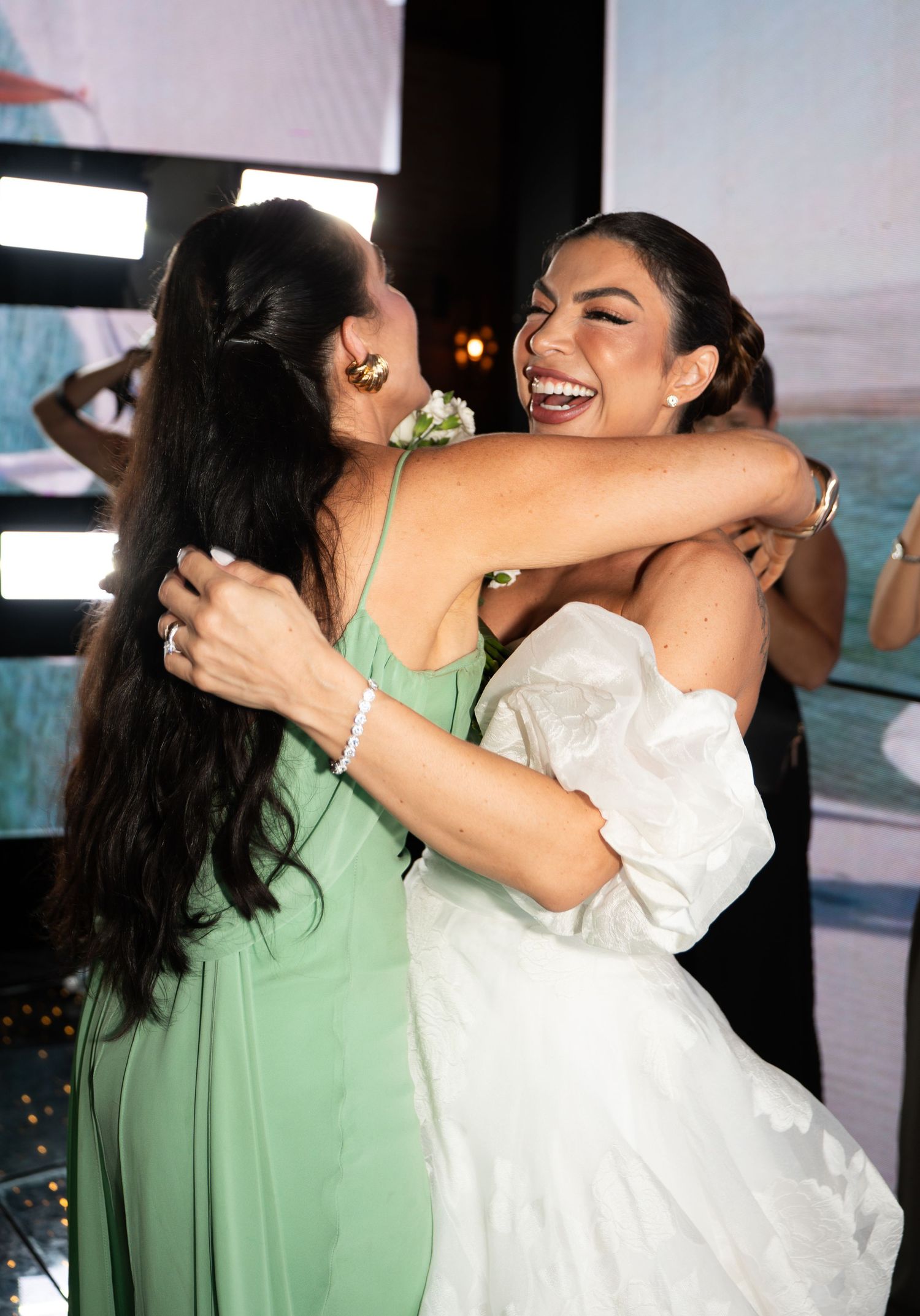 Two women share a joyful hug at a formal event, one in a green dress and one in white.