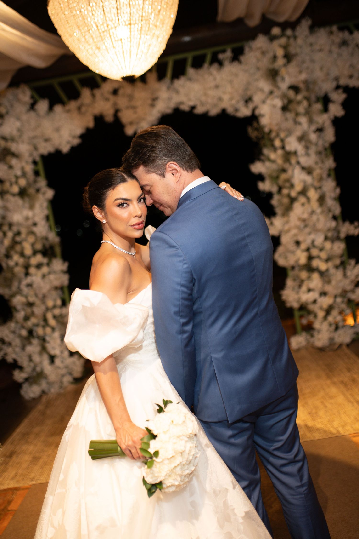 Bride in white off-shoulder gown holding bouquet embraces groom in blue suit before floral arch at elegant wedding reception.