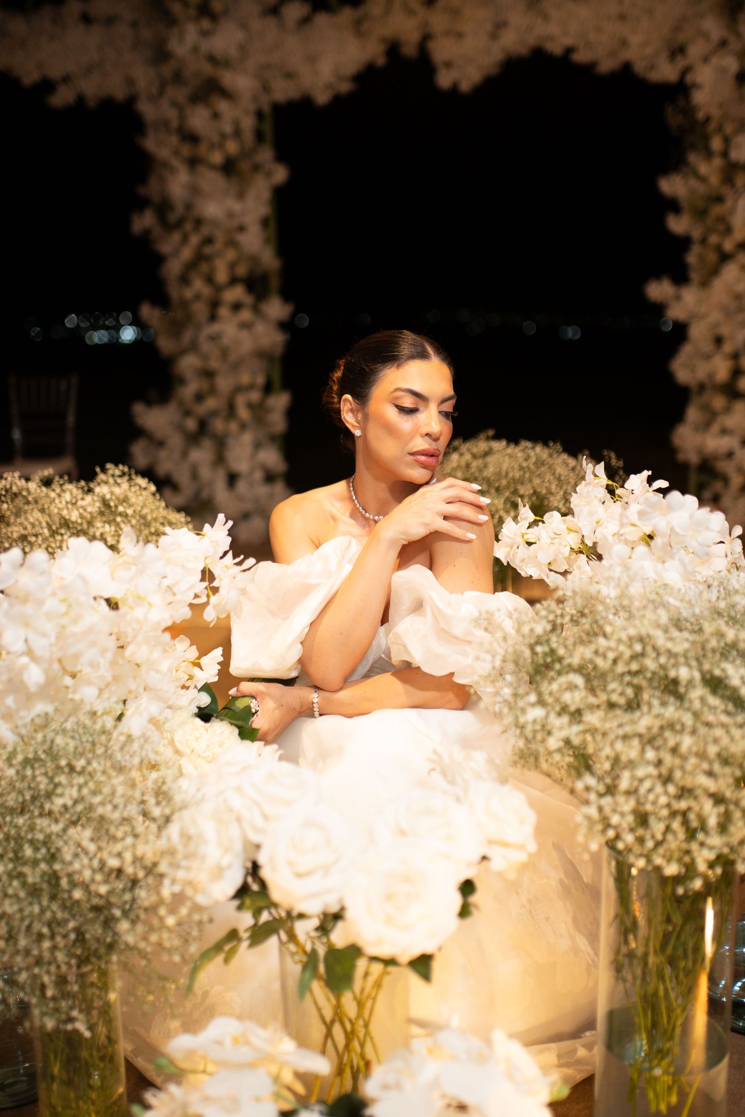 Elegant bride in white off-shoulder gown surrounded by lush white floral arrangements at a nighttime wedding celebration.