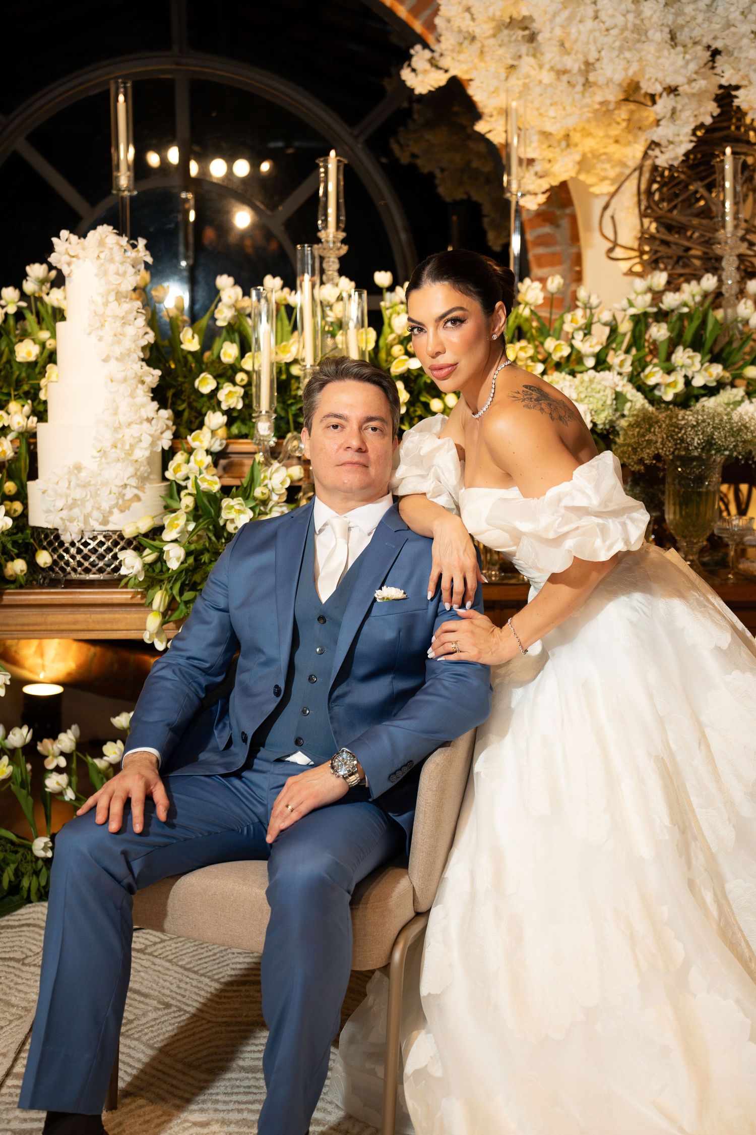 Elegant wedding couple posing before floral backdrop, groom in blue suit, bride in white off-shoulder gown.