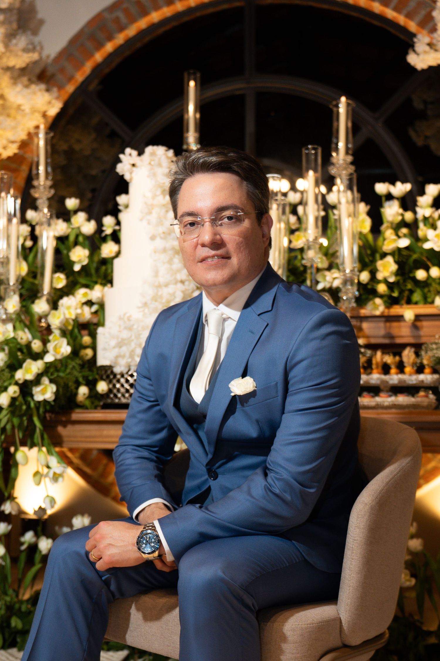 Man in blue suit seated before wedding cake and floral arrangement at elegant reception venue.