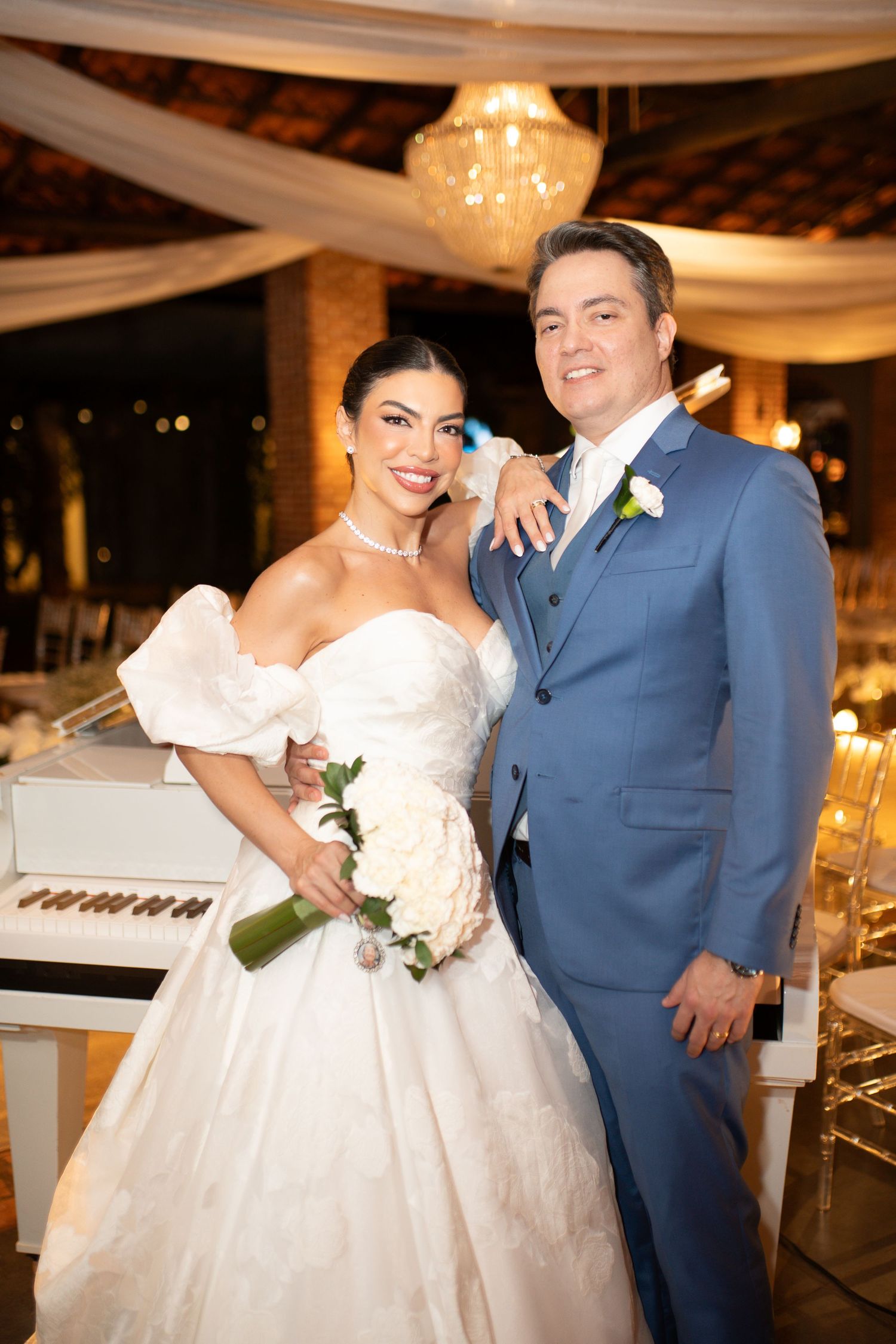 A bride in a white ball gown holding roses poses with a groom in a blue suit at an elegant wedding reception.