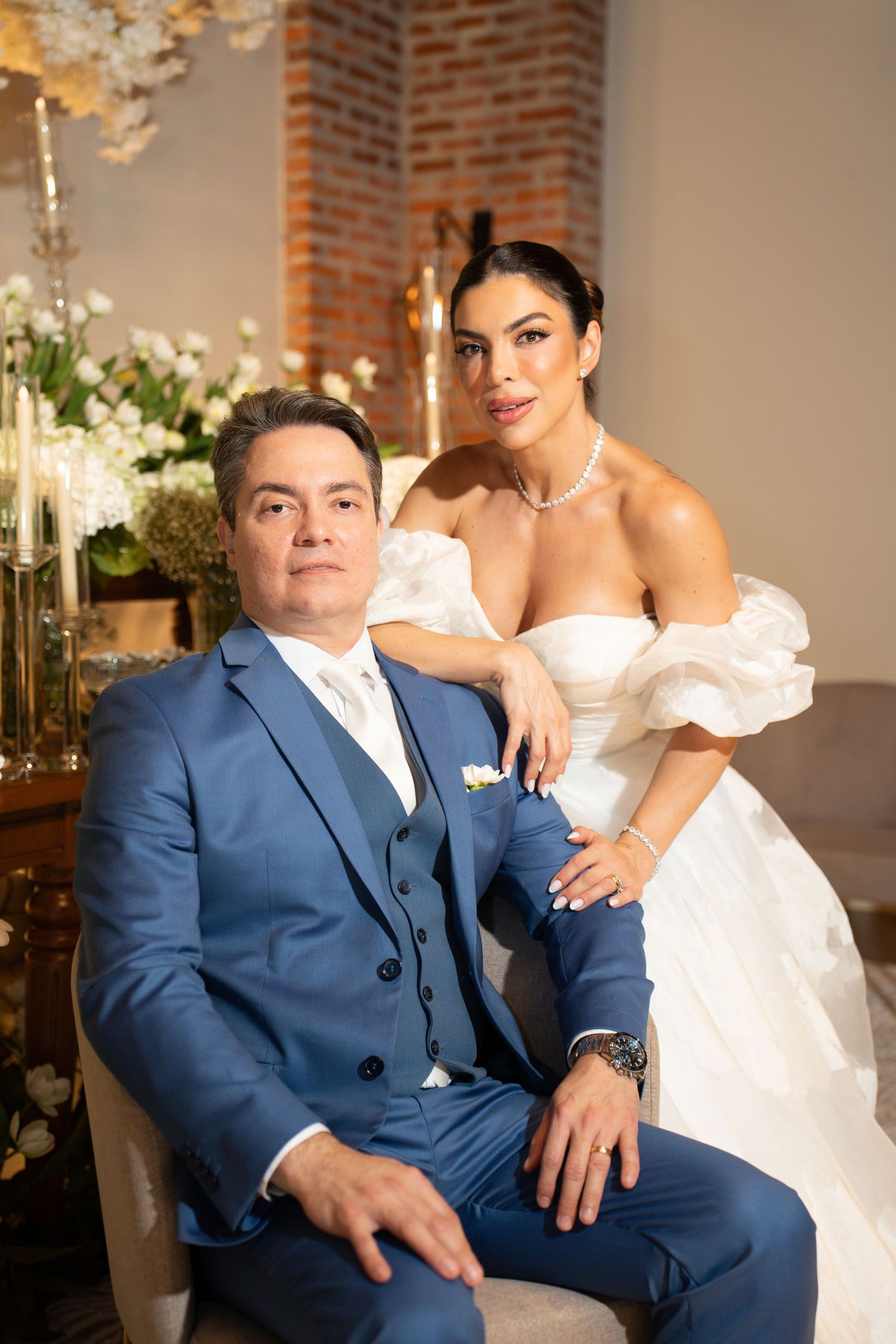 Elegant bride in white off-shoulder gown stands behind groom in blue suit, posing for formal wedding portrait indoors.