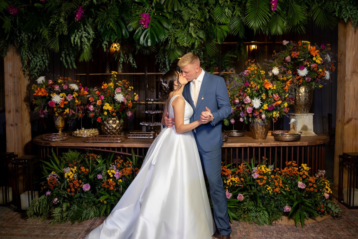 Bride and groom share a romantic kiss surrounded by lush tropical greenery and colorful floral arrangements.