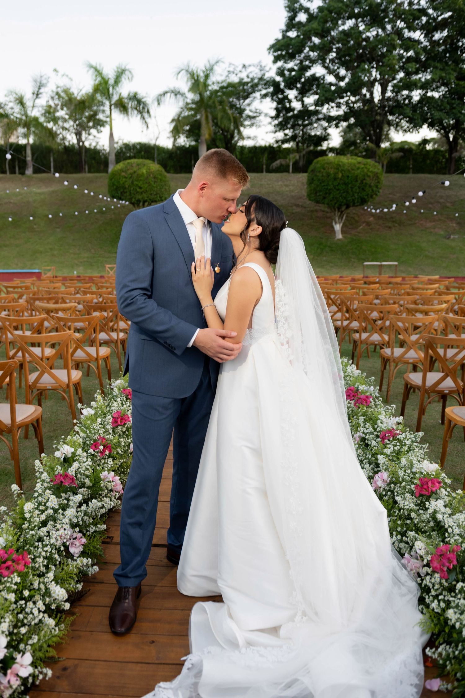 Newlyweds kiss at outdoor wedding altar lined with pink and white flowers, wooden chairs, and string lights.