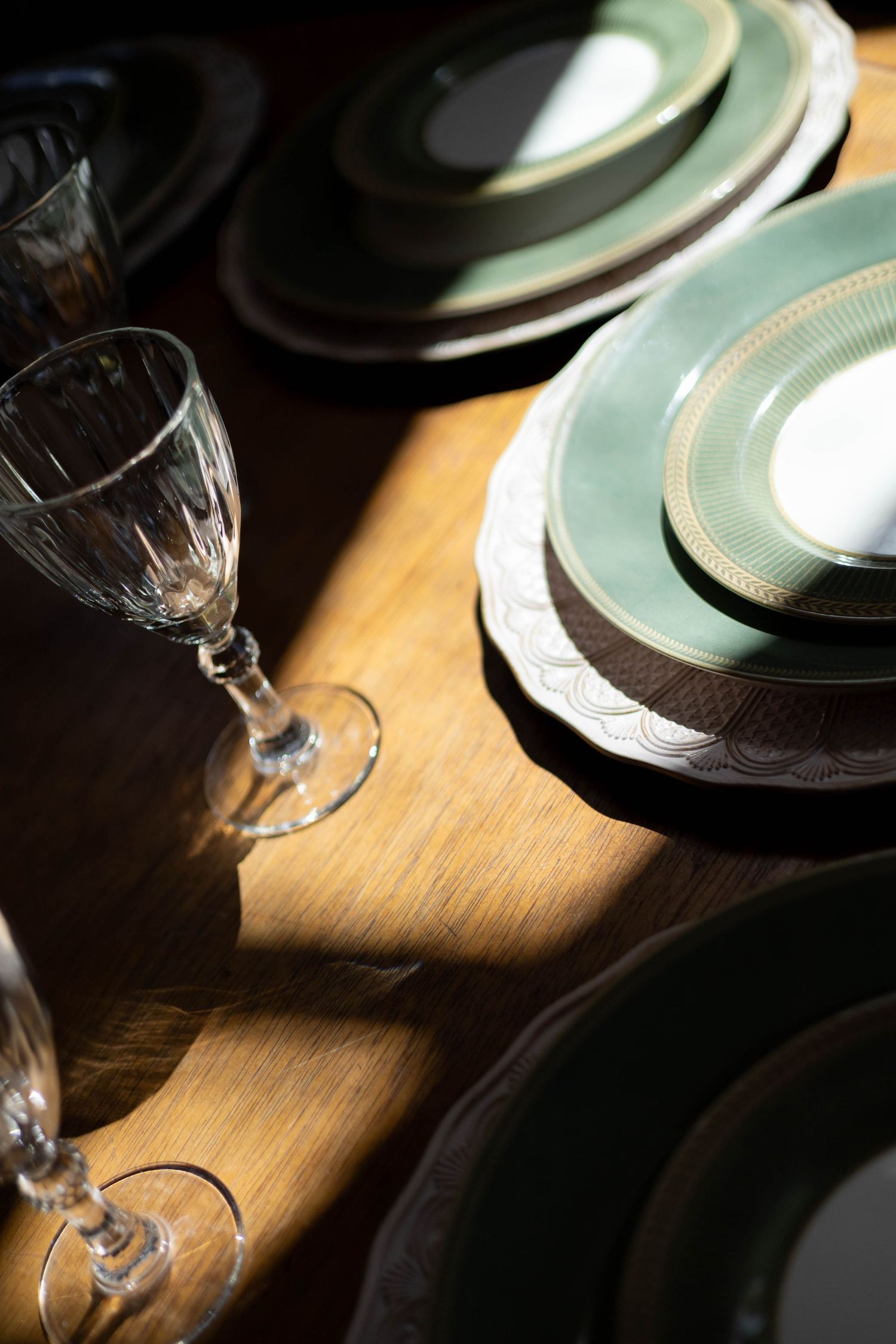 Elegant table setting with stacked green and gold rimmed plates and crystal wine glasses on a wooden table in soft light.