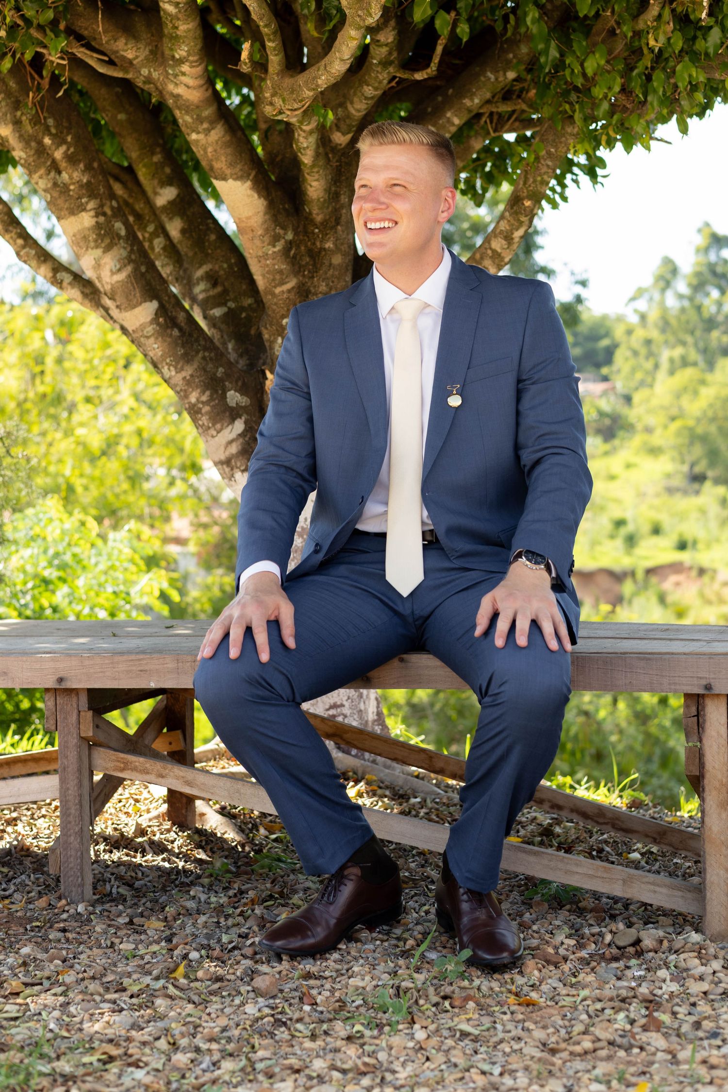 Smiling man in navy blue suit sitting on wooden bench outdoors under a tree on a sunny day.