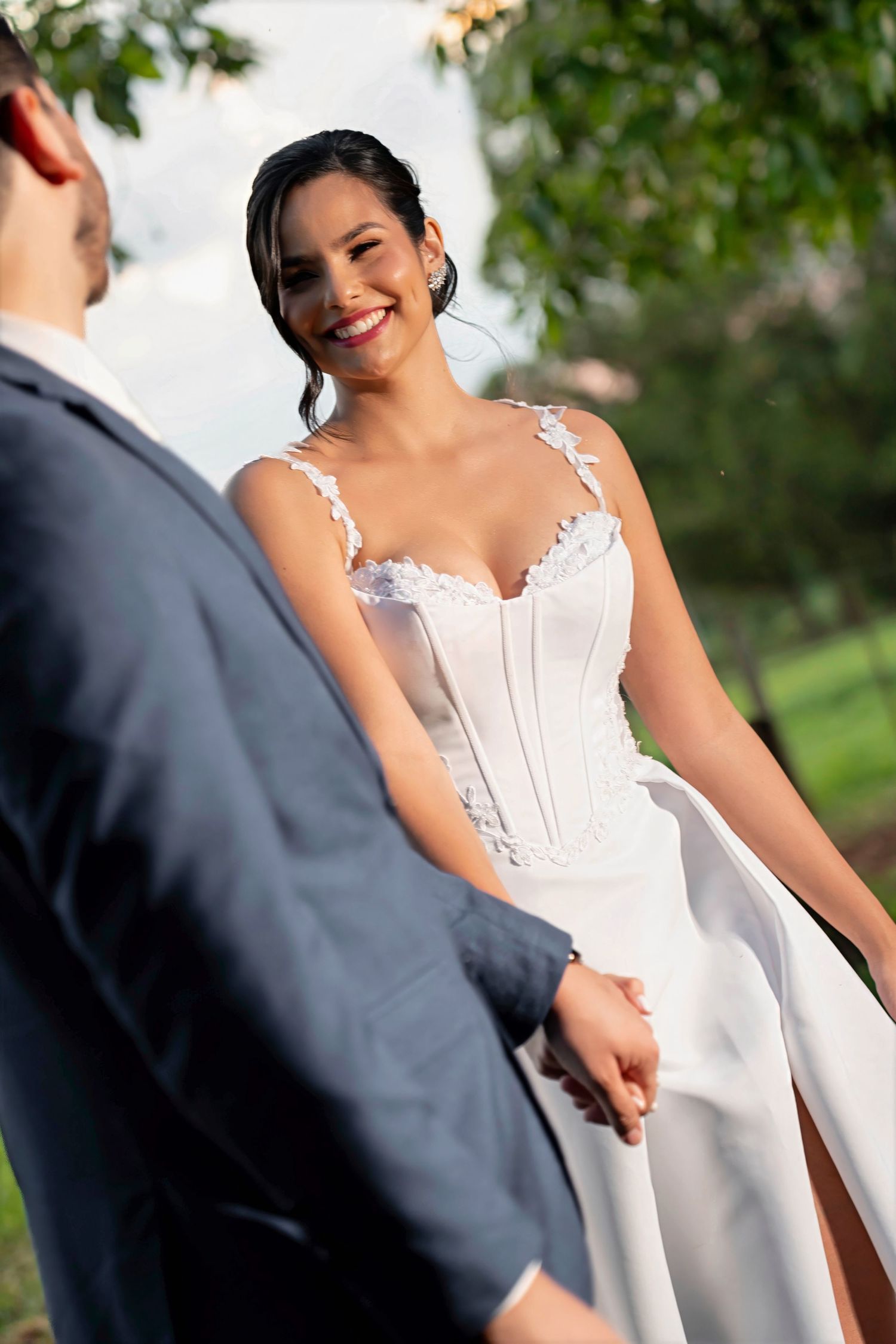 Smiling bride in white lace gown holds hands with groom outdoors during romantic wedding ceremony.