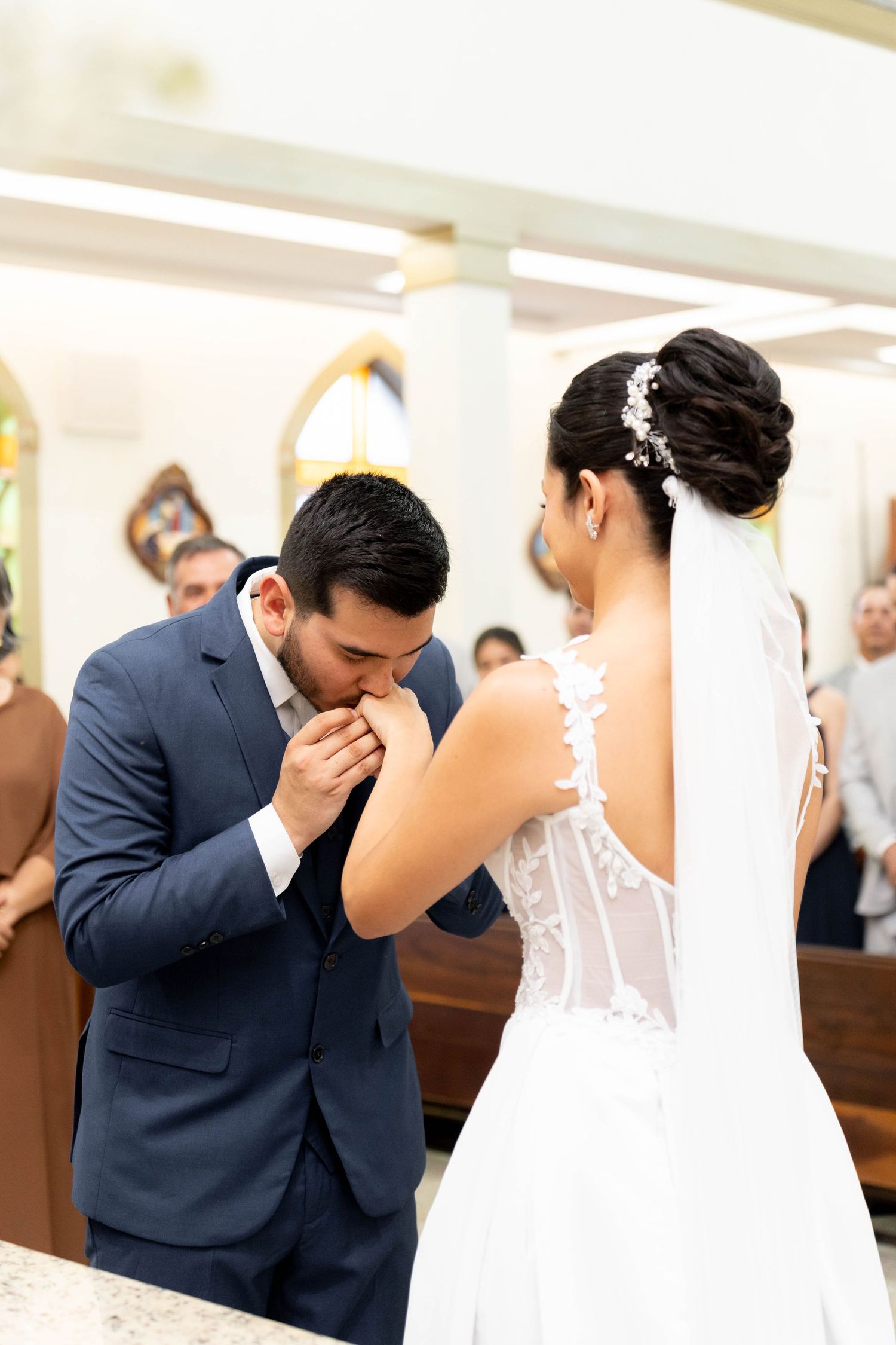 Groom in blue suit kisses bride's hand during wedding ceremony in church.