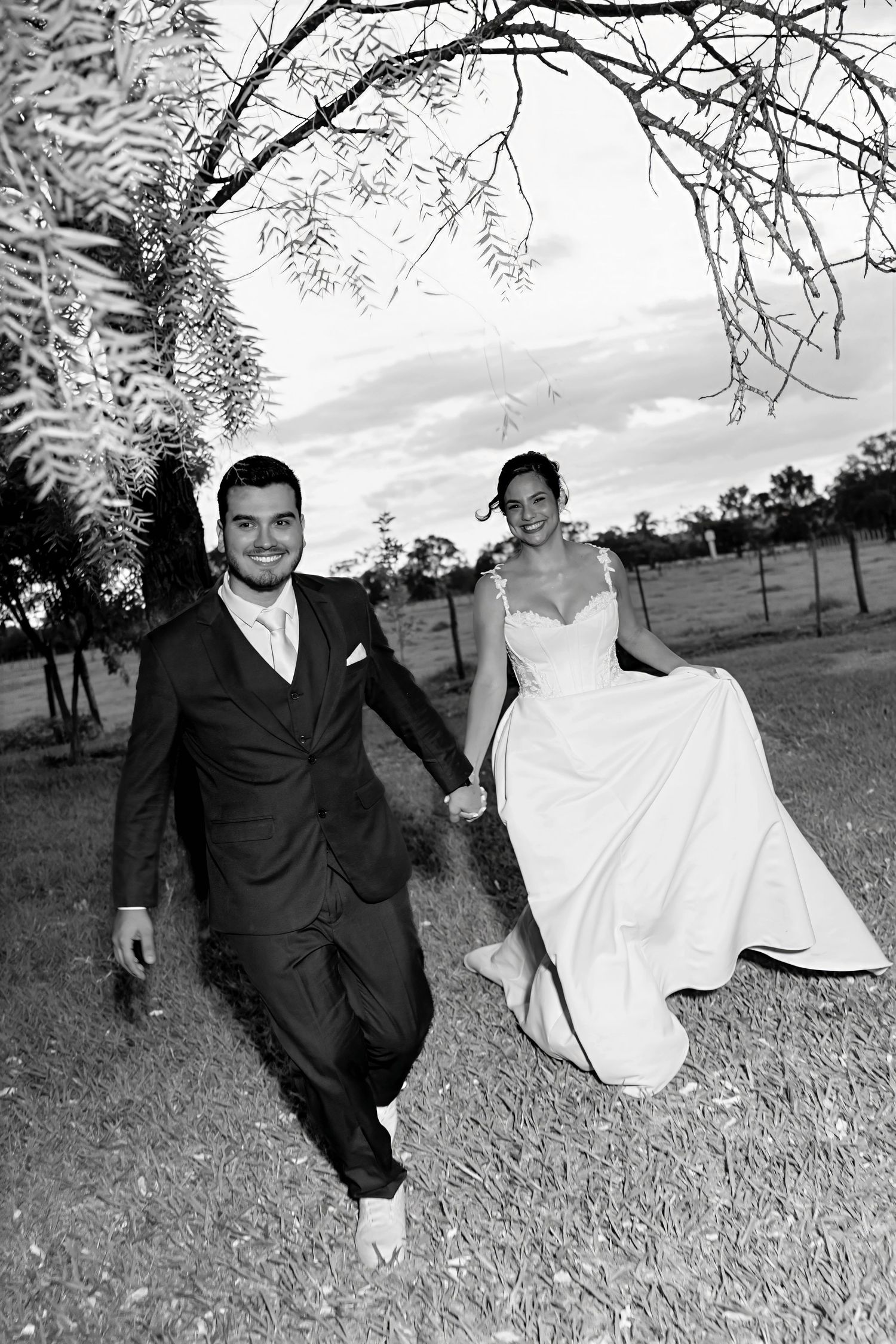Black and white photo of a smiling groom in a suit leading his bride in a white gown by the hand outdoors.
