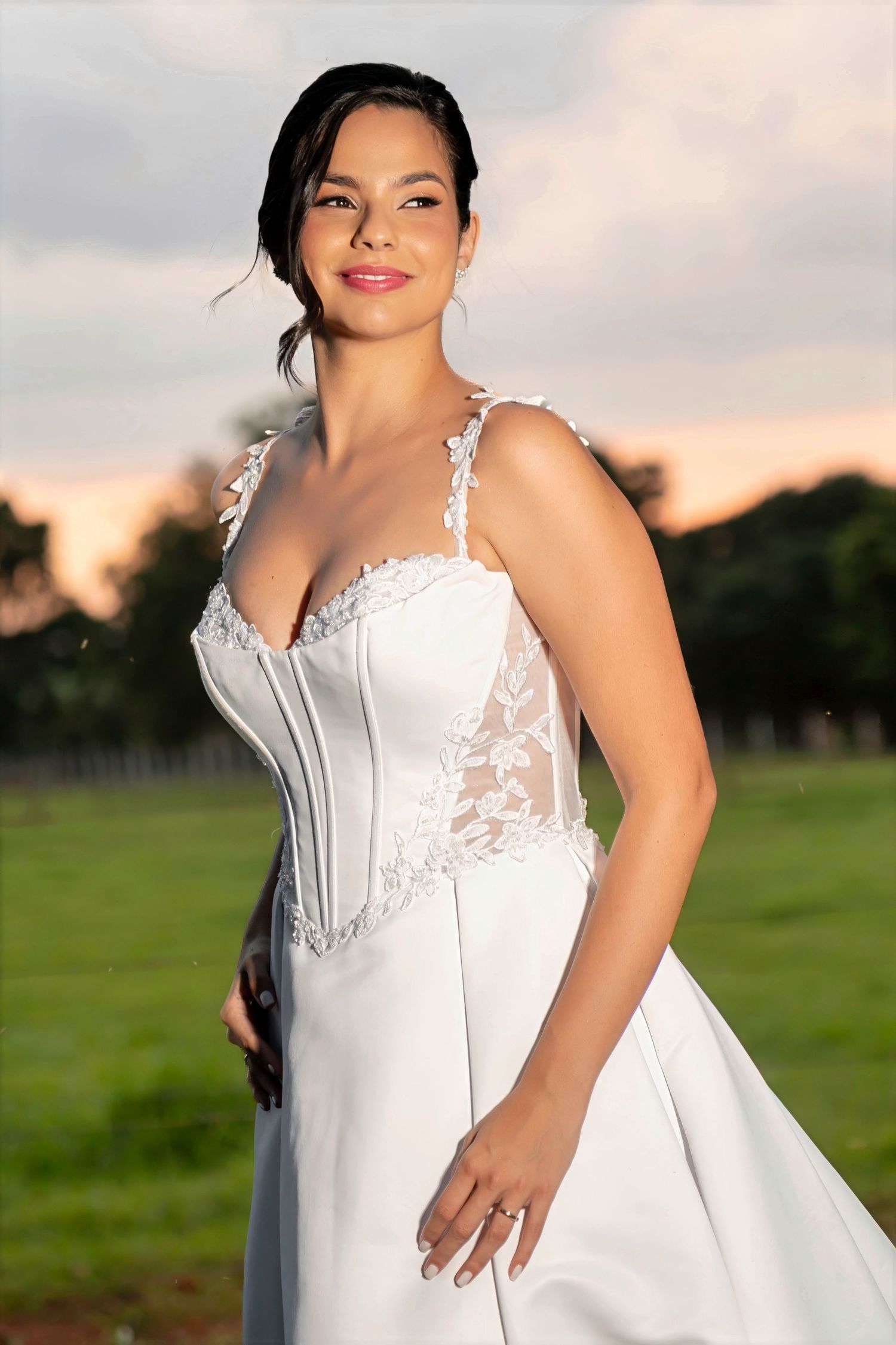 Bride in elegant white lace corset wedding gown posing outdoors at sunset with green landscape background.