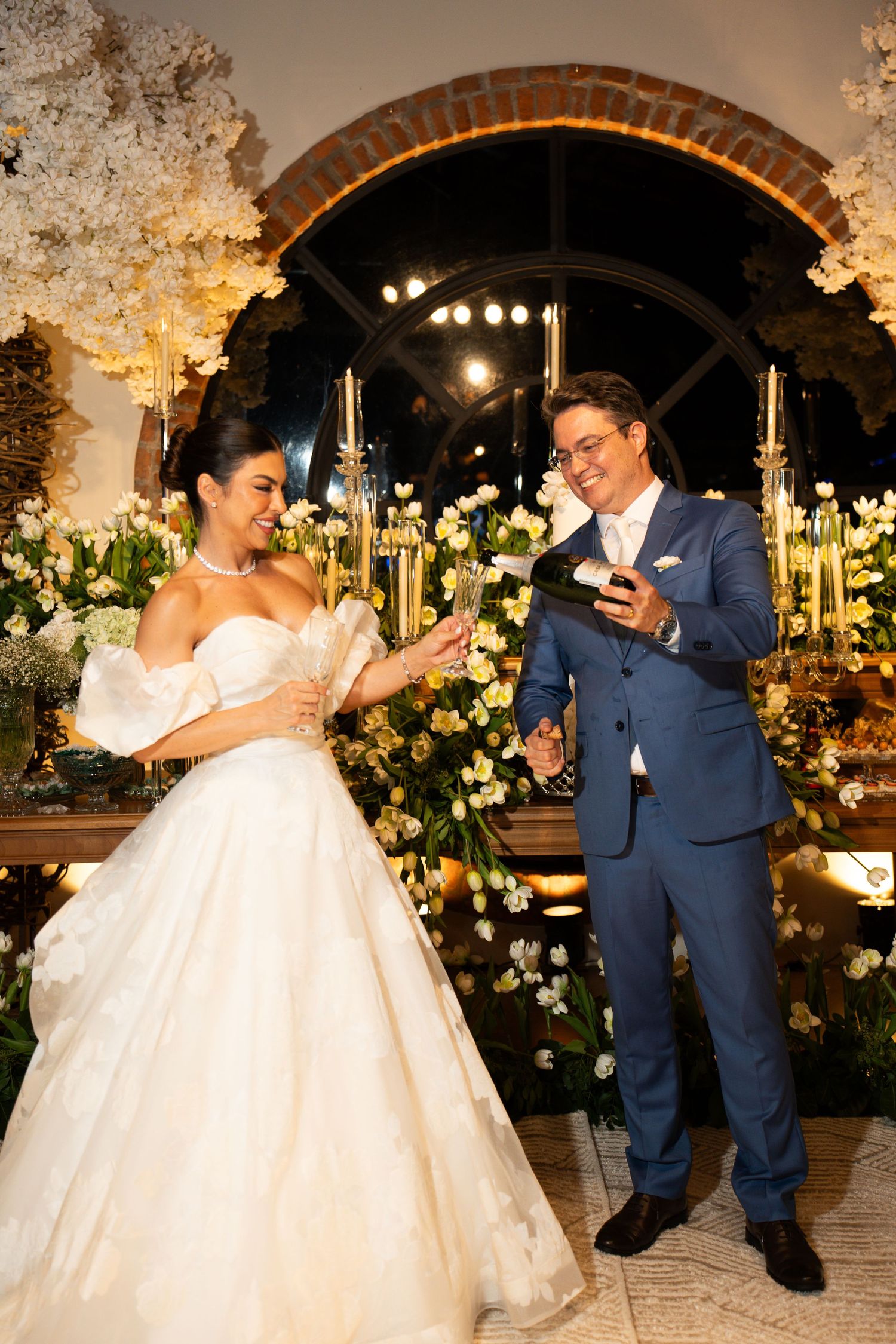 Bride in white ball gown and groom in blue suit pouring champagne at elegant floral wedding reception.