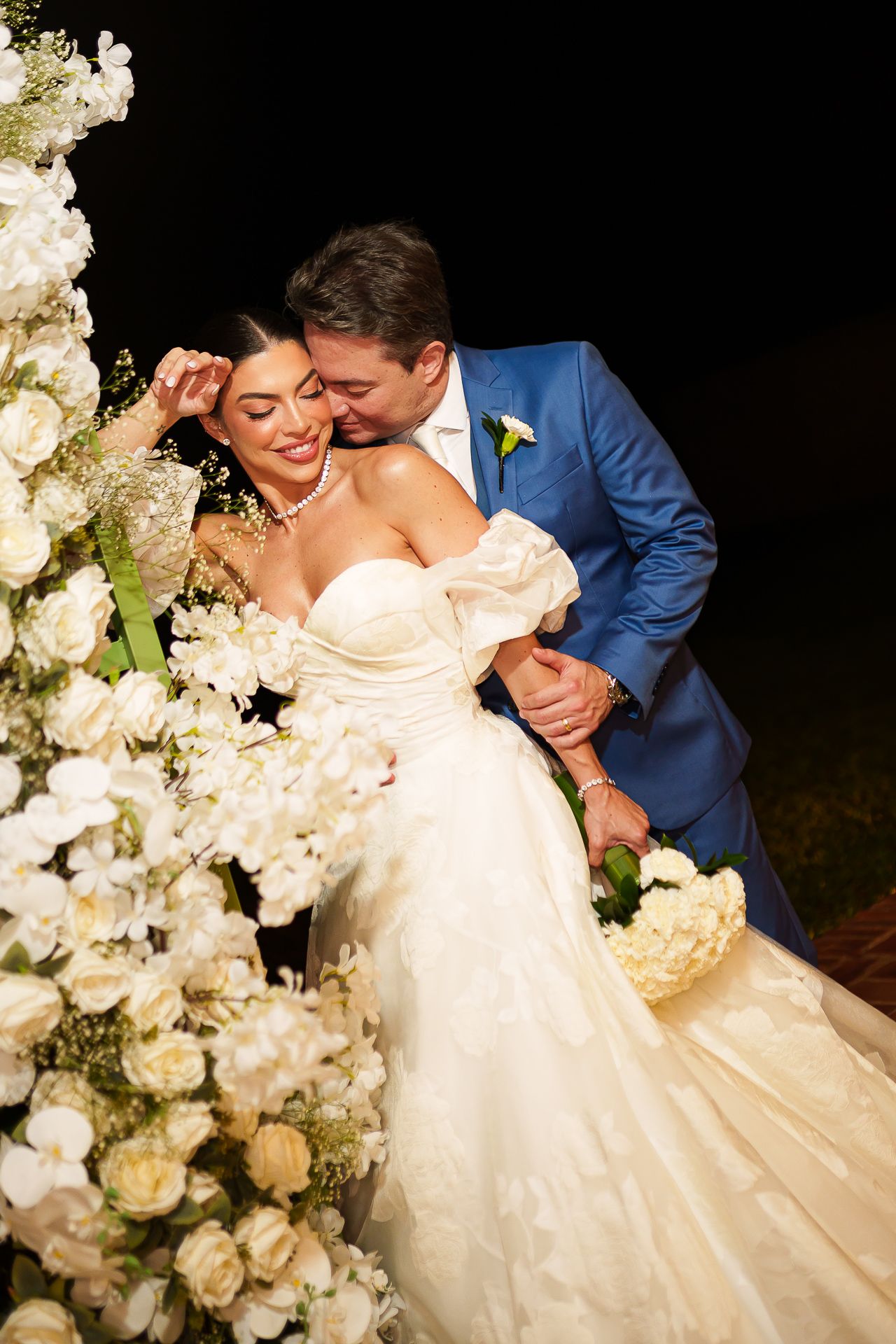 Bride in off-shoulder gown and groom in blue suit share a romantic kiss beside white floral arrangements at night wedding.