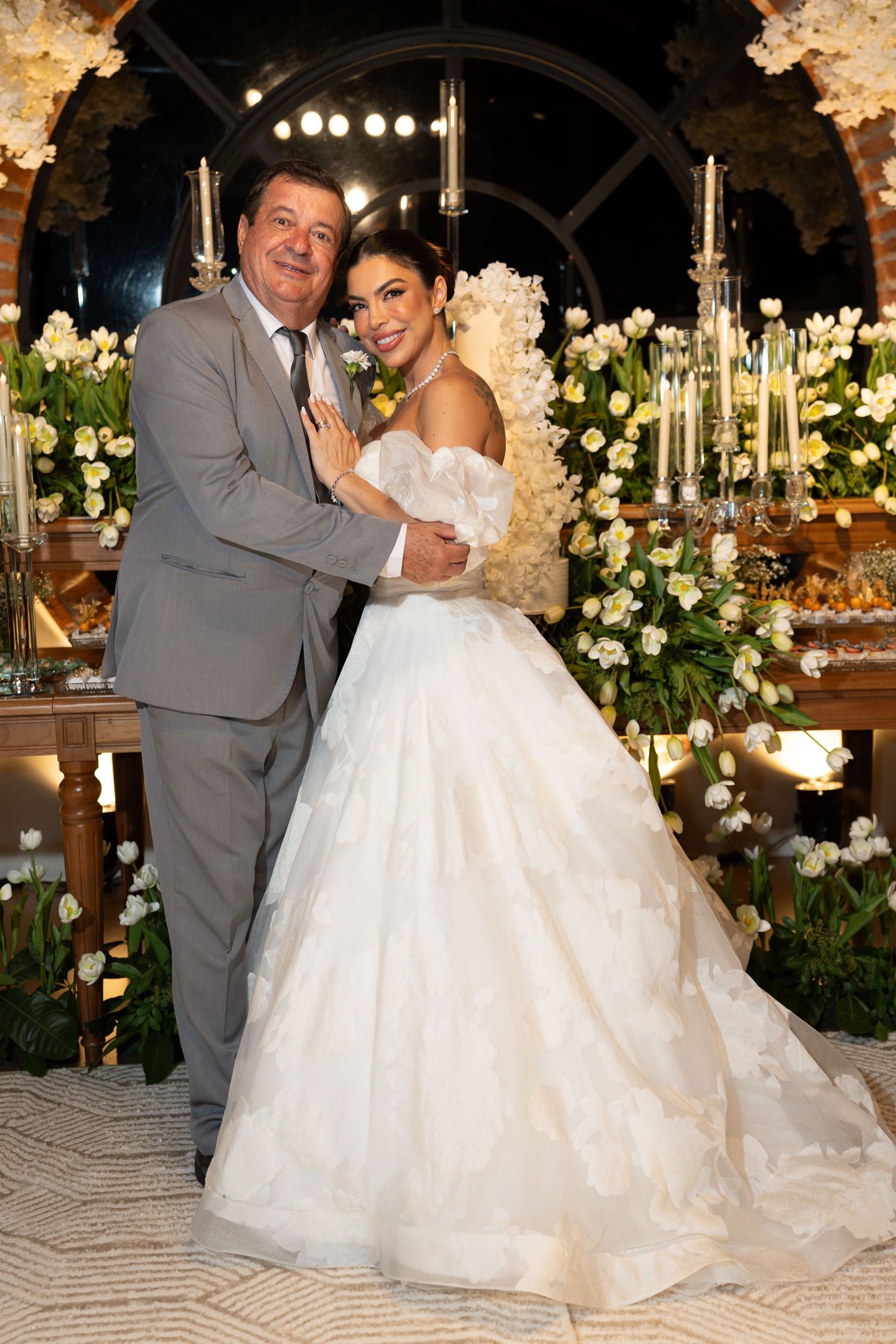 Bride in white ball gown and groom in gray suit pose together at elegant floral wedding reception.