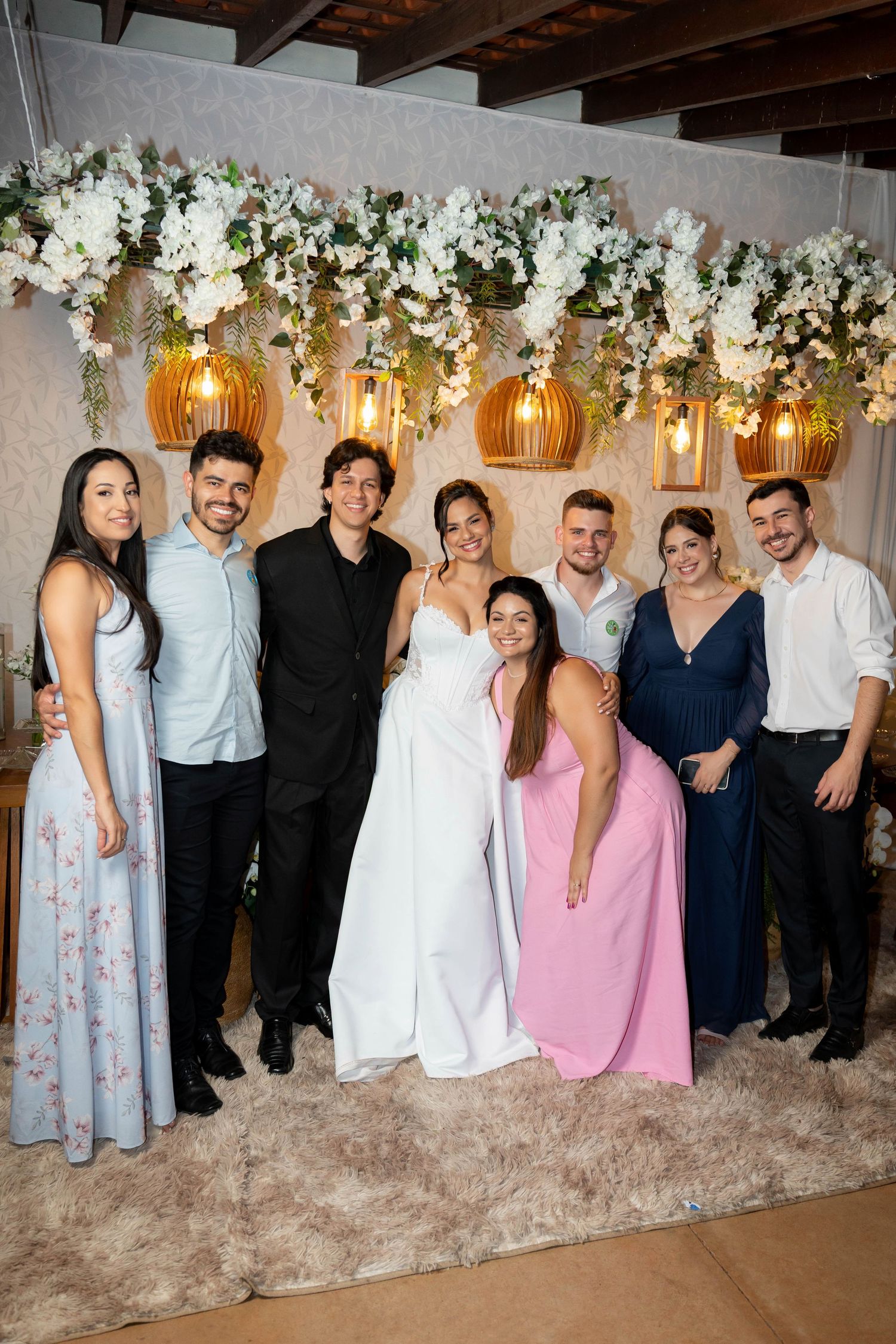 A joyful wedding group photo with bride in white gown, groom, and guests posing under floral decor with warm hanging lights.
