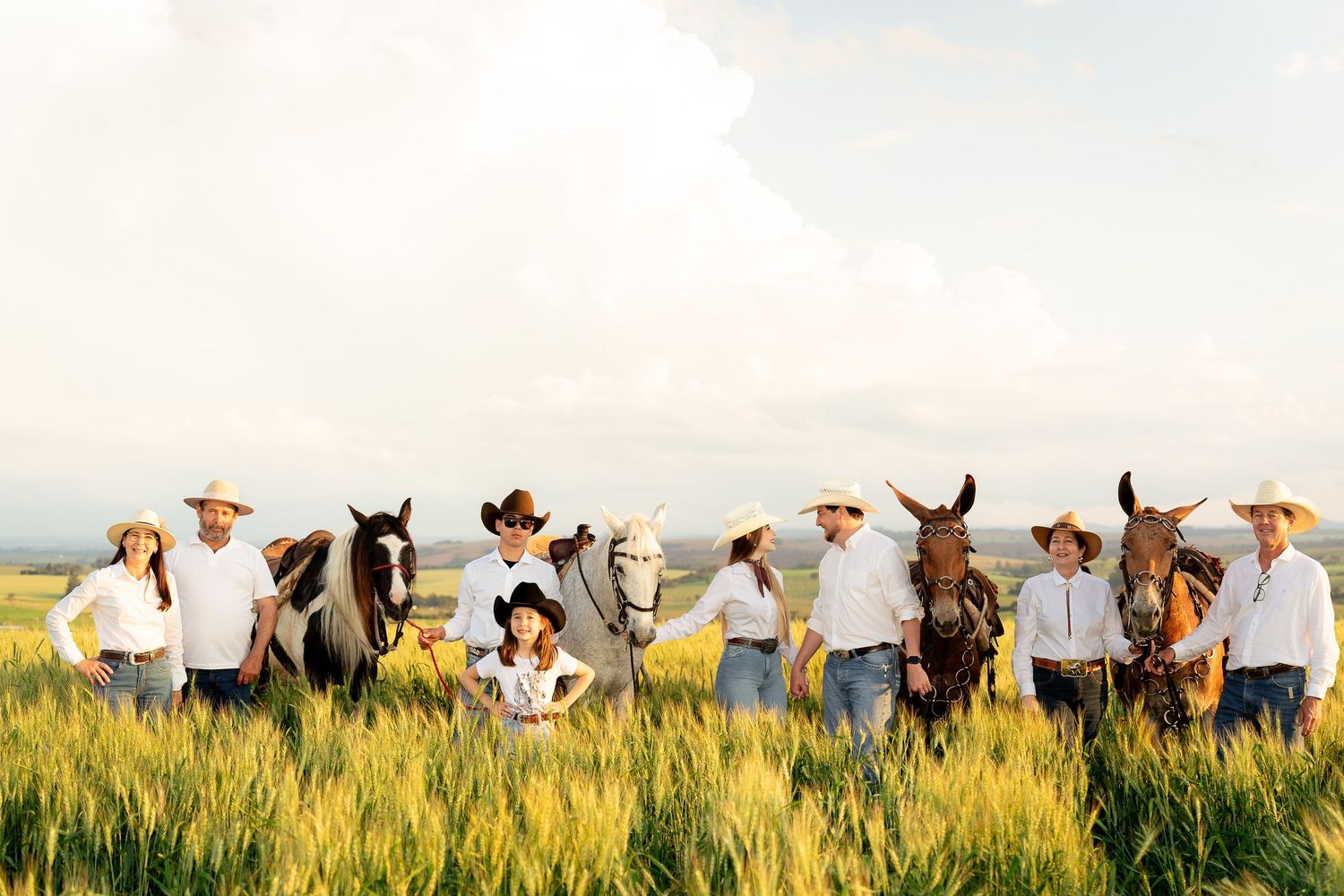 A group of people in cowboy hats stand with horses and mules in a lush green field under a cloudy sky.