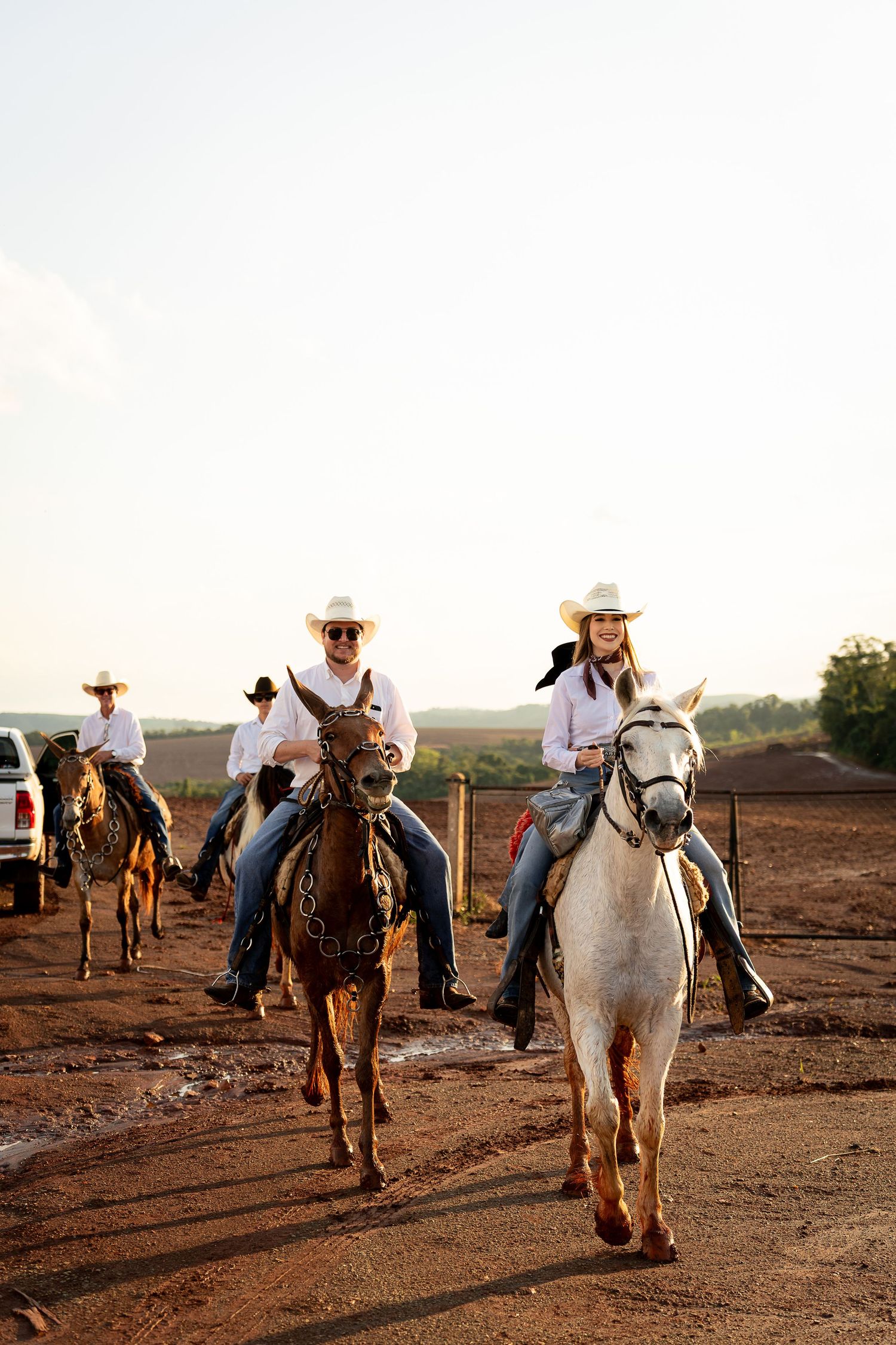 Cowboys and cowgirls in white hats ride horses along a dirt path at golden hour on a ranch.