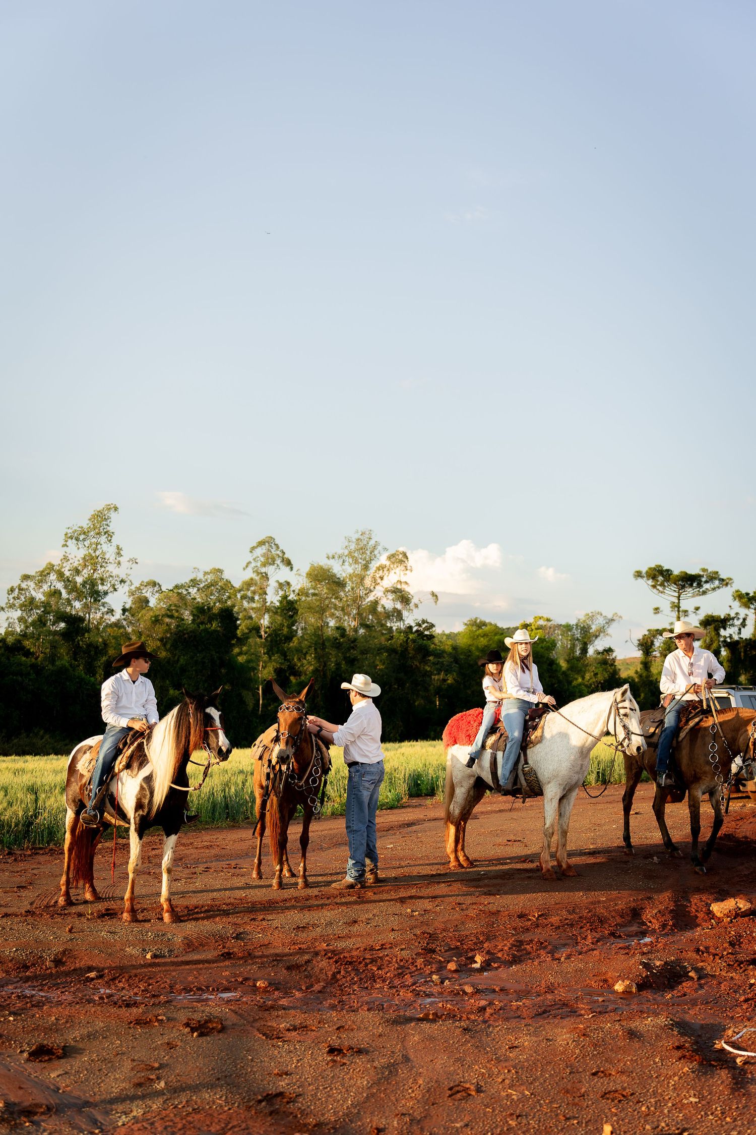 Cowboys on horseback gather on a dirt path beside green fields during golden hour in a rural countryside setting.